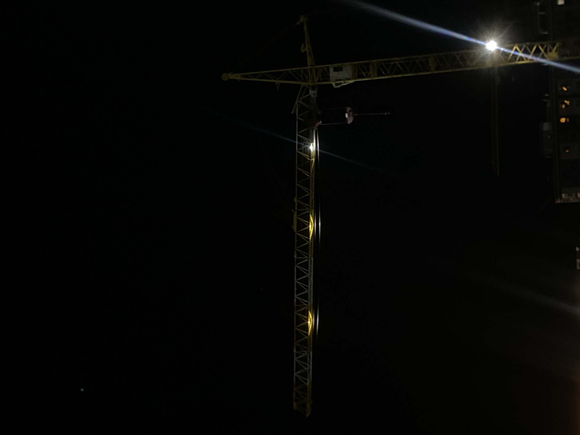 A construction crane is illuminated against a dark night sky. The crane is yellow and consists of a tall vertical tower and a long horizontal jib. Lights are visible along the jib, casting a yellow glow. A hook hangs from the jib. In the background, faint lights from buildings suggest a city environment. The image was captured in Yangi Andijan, Uzbekistan.