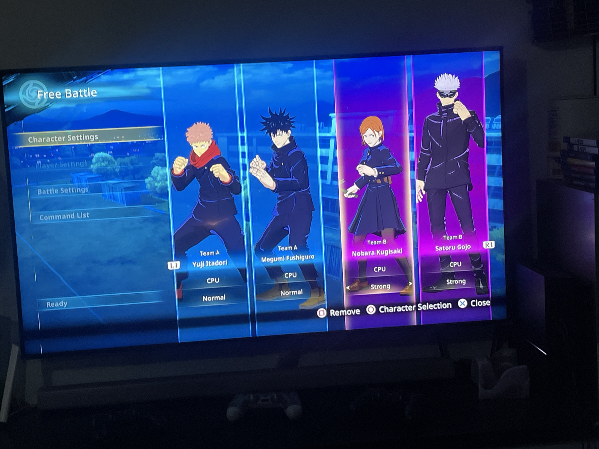 The image shows a TV screen displaying the character selection screen of a fighting game set in "Free Battle" mode. On the left side of the screen, a menu is visible with options such as "Character Settings," "Player Settings," "Battle Settings," and "Command List." Four characters are selected for the teams: Yuji Itadori and Megumi Fushiguro for Team A, and Nobara Kugisaki and Satoru Gojo for Team B. All four characters are controlled by the CPU, with Yuji Itadori and Megumi Fushiguro set to "Normal" difficulty, while Nobara Kugisaki and Satoru Gojo are set to "Strong" difficulty.
The bottom of the screen displays options such as "Remove," "Character Selection," and "Close."
The location of Ado, Nigeria, is not related to the contents of the image.