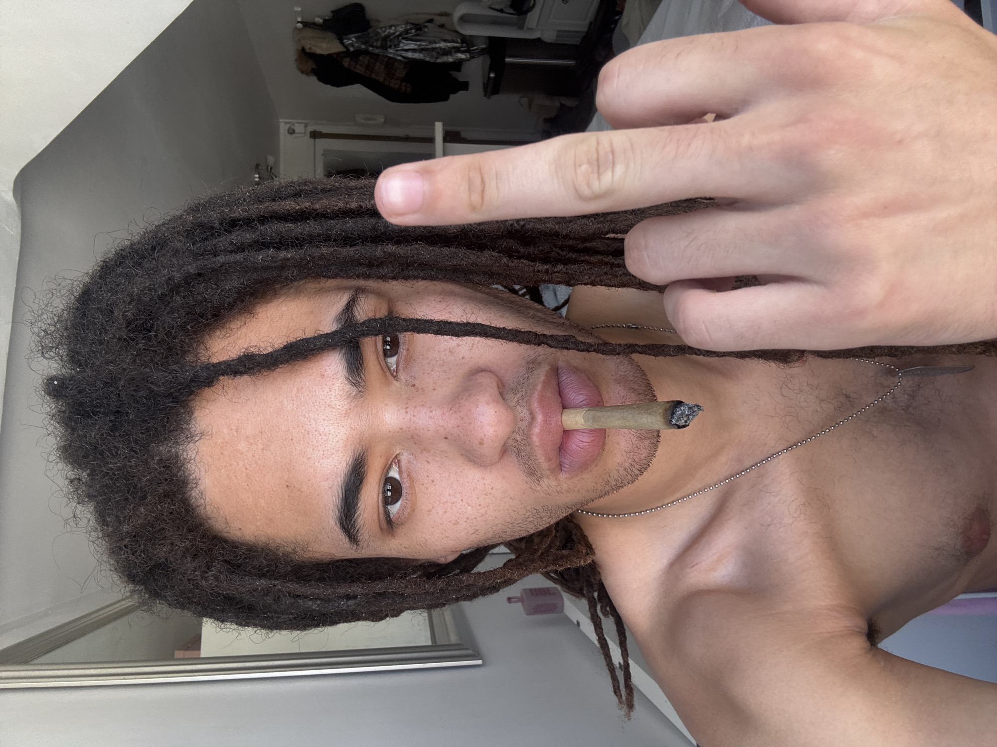A person with dark skin and brown dreadlocks is holding a lit cigarette or joint in their mouth. The person's upper body is visible, revealing a bare chest and a silver chain necklace. The person is giving the middle finger with their right hand. The background appears to be an indoor setting, possibly a room with a door and a glimpse of clothing or other items in the distance. There is no indication of the location being Paris, France, or any other specific place.