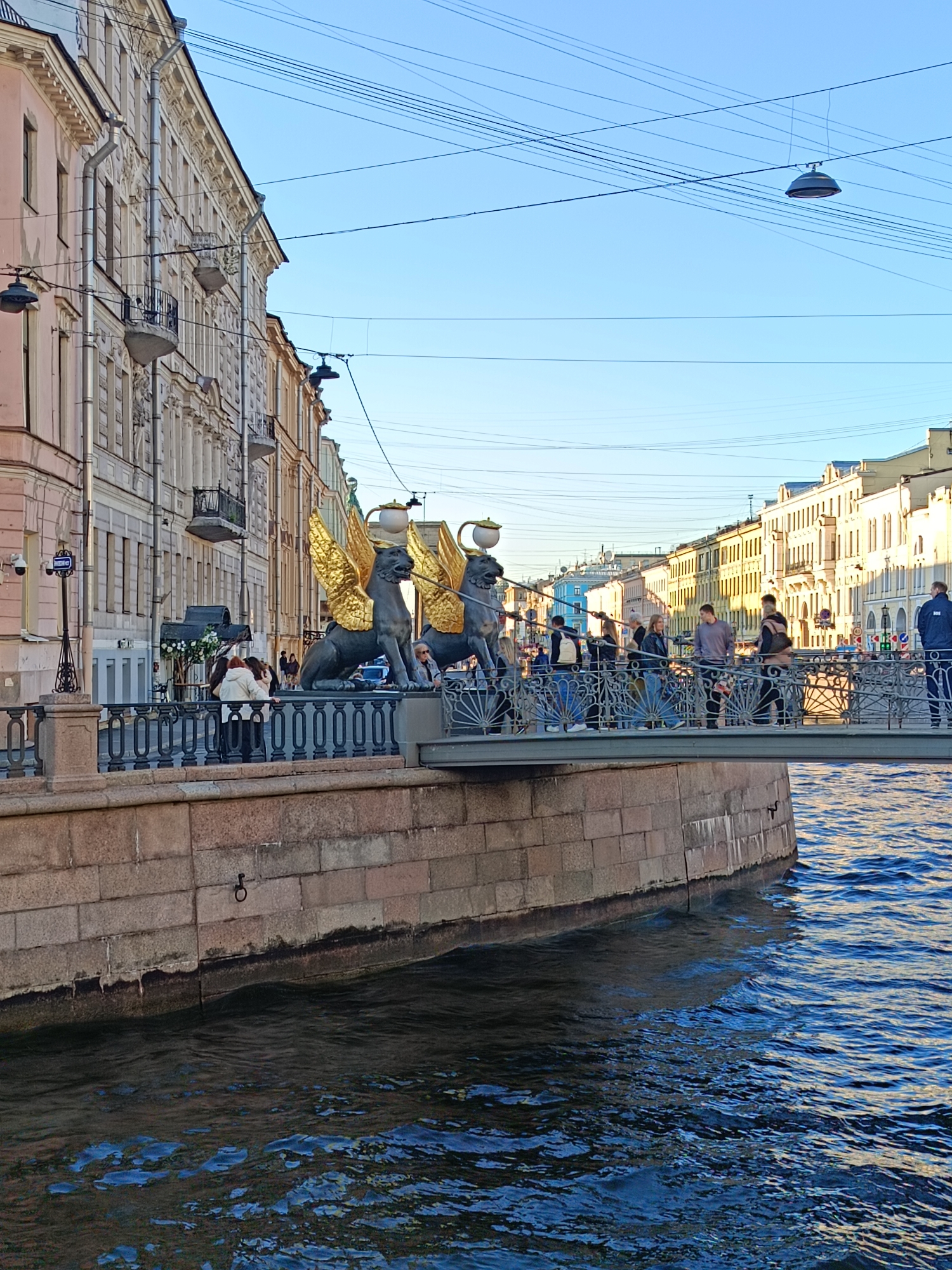 The image captures a vibrant scene on the Bank Bridge in Saint Petersburg, Russia. The bridge spans over a body of water, reflecting a deep blue with visible ripples and movement. The bridge is lined with people, casually dressed, some strolling and others pausing to enjoy the view.
The bridge's parapets feature intricate metalwork, supporting the iconic statues of griffins with gilded wings. These mythological creatures stand sentinel, facing outward with their heads held high.
On the left, a building with ornate facades and balconies adds architectural interest. The sky above is a clear, pale blue, providing a bright backdrop to the scene. Overhead, there are several electrical wires and one lamppost.
