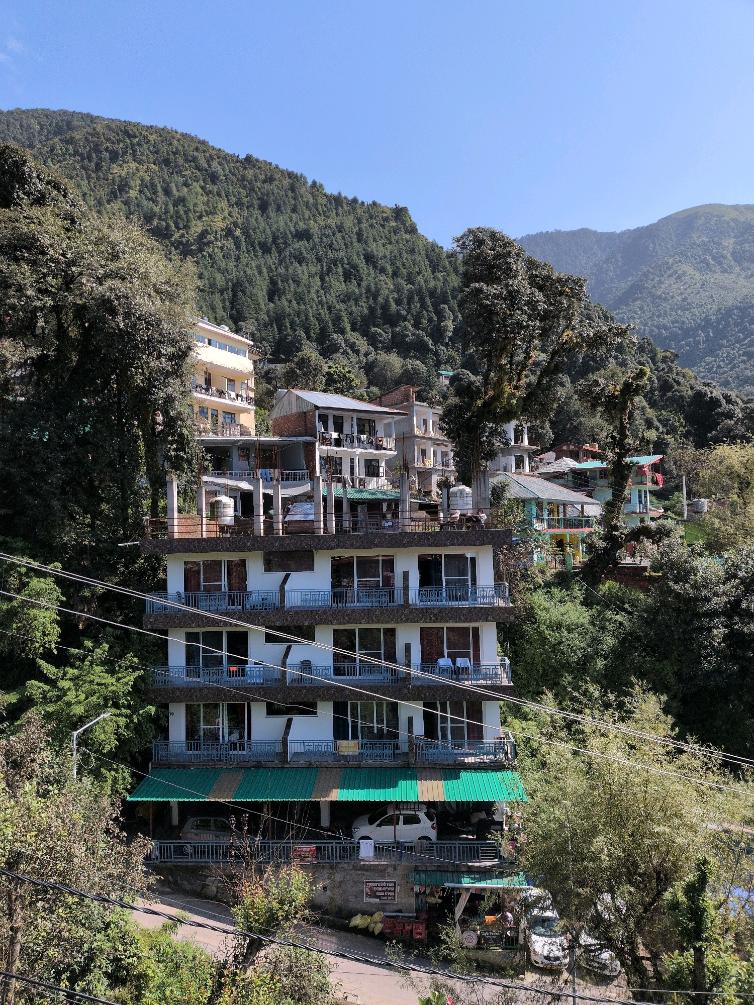 The image shows a multi-story building nestled on a steep, tree-covered hillside in Bhagsu, India. The building, primarily white with brown accents and numerous balconies, appears to be a hotel or guesthouse. In front of the building, several cars are parked, and there are awnings covering some of the lower levels, suggesting shops or restaurants. The surrounding environment is lush with dense green vegetation, characteristic of the Himalayan foothills. The sky is a clear blue, indicating fair weather. The scene depicts a typical mountain town setting with a mix of natural landscape and man-made structures.