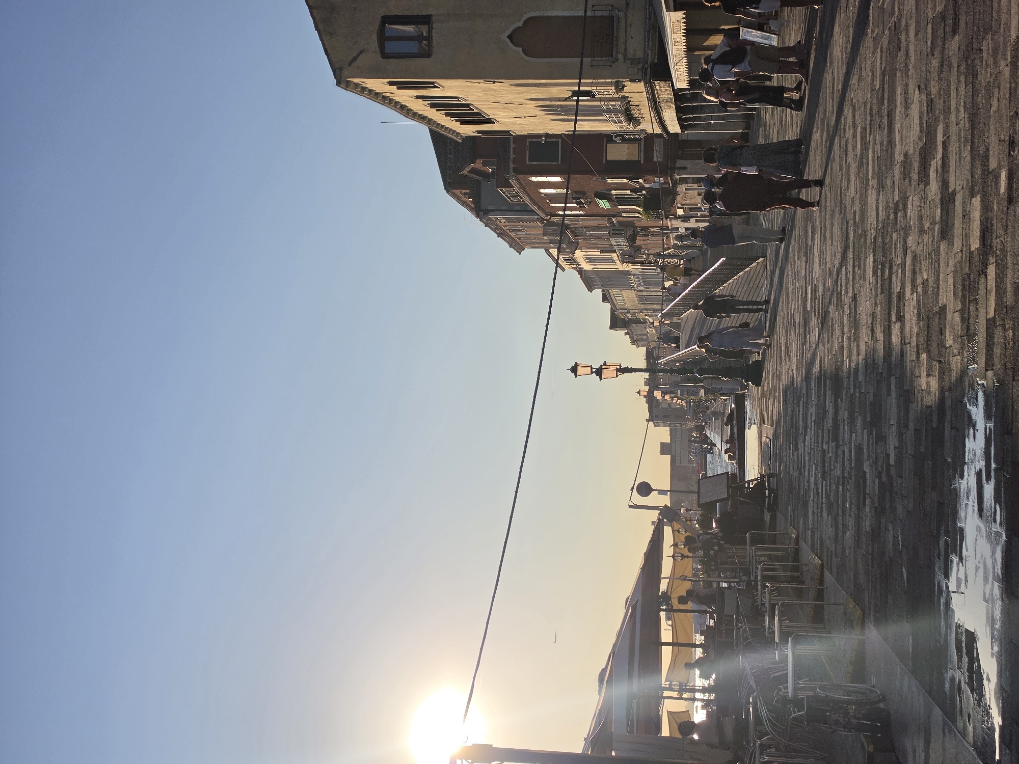 The image depicts a sun-drenched scene along a paved walkway in Venice, Italy, likely during late afternoon or early evening. The sun is low in the sky, casting long shadows and creating a bright glare on the left side of the image, partially obscuring the view of umbrellas and tables belonging to an outdoor seating area of a restaurant or cafe.
Several people are visible walking along the walkway and sitting at tables. On the left, silhouettes of people are seen behind cafe umbrellas. Further down the walkway, a group of individuals are walking in various directions, some engaging in conversation. There are architectural elements typical of Venice, with multi-story buildings lining the right side of the walkway. These buildings feature numerous windows and decorative architectural details. Lamp posts are strategically placed along the walkway, adding to the ambiance. The ground is paved with cobblestones, some of which are wet, reflecting the sunlight. In the distance, a bridge can be seen, with people walking across it. The sky is clear and a pale blue, indicating good weather. The overall impression is one of a pleasant, active urban environment in Venice.