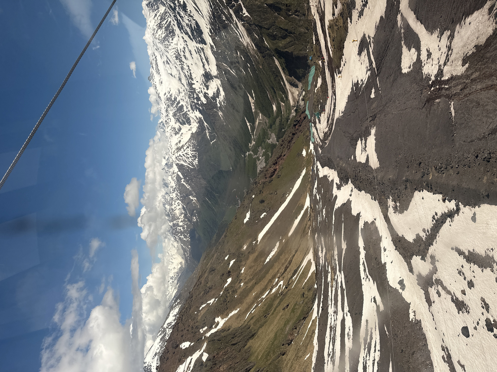 The image captures a panoramic view of a mountainous landscape, likely taken from a cable car, with snow-capped peaks in the background under a bright blue sky with scattered clouds. In the foreground, a ski slope is visible, partially covered in snow and dark volcanic rock or soil. The slope is uneven, with patches of white snow interspersed with dark earth and rocks. Further down the mountain, a valley unfolds with a winding river or road and areas of green vegetation. Small structures and what appear to be turquoise-colored pools are visible in the mid-ground. The overall impression is of a natural, rugged terrain with elements of human infrastructure. The location is identified as Terskol, Russia, suggesting a ski resort or mountainous region in that area.
