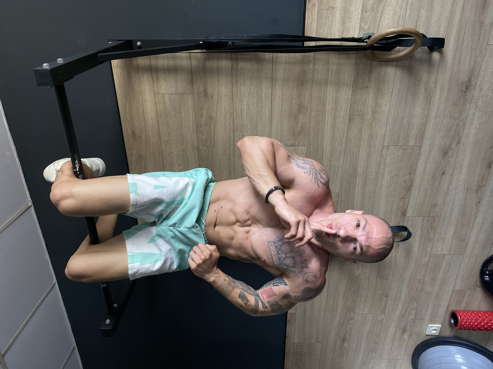 A man with a shaved head and tattoos on his arms and torso stands in a room with light wood flooring and a black wall. He wears white and turquoise patterned shorts and white shoes, positioned near a black metal pull-up bar and hanging gymnastic rings. The man is gesturing towards his mouth with his right index finger. Other objects in the scene include a red foam roller and a gray exercise ball. The location cannot be determined from the image alone.
