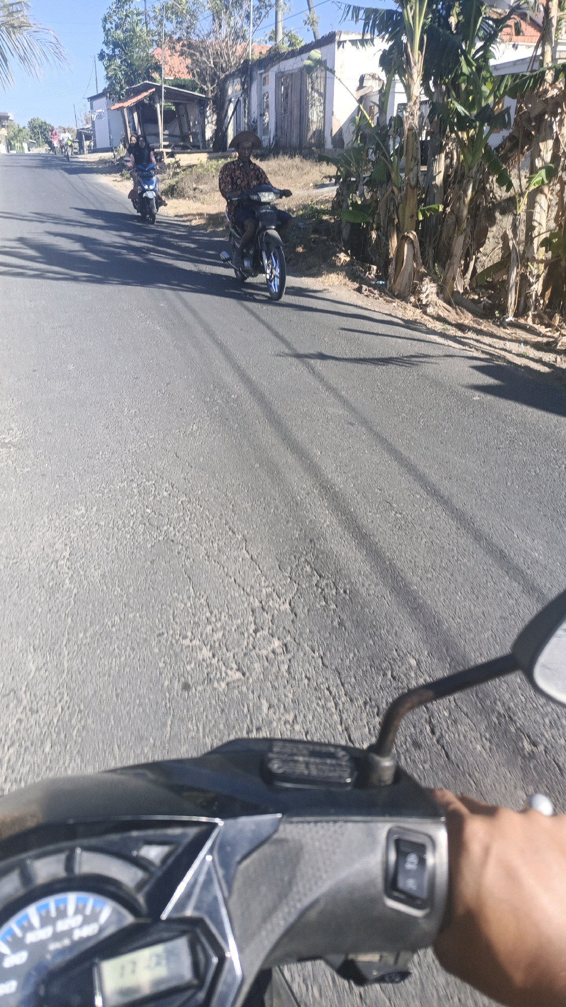 The image depicts a sunny day on a rural road in Gayam, Indonesia. The perspective is from the viewpoint of someone riding a motorcycle, as indicated by the handlebars and dashboard in the foreground. The road is asphalt and shows some wear and tear. Two motorcycles are visible in the mid-ground. The first motorcycle, closer to the viewer, is ridden by an individual wearing a patterned shirt and a wide-brimmed hat, heading towards the viewer. The second motorcycle, further down the road, carries two passengers, a driver and a person behind, also moving away from the viewer. The background features a row of buildings with weathered facades, some with visible damage, and lush green vegetation, including banana trees. The overall scene suggests a typical day on a country road in a tropical climate.