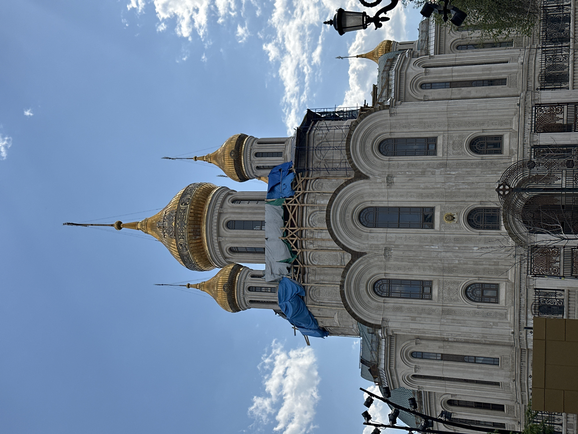 The image shows an upward, oblique view of the Cathedral of Christ the Saviour in Moscow, Russia. The cathedral features multiple gold-domed cupolas rising against a bright blue sky with scattered white clouds. Part of the cathedral's facade is visible, showcasing its arched windows and ornate carvings. Scaffolding and blue tarpaulins are present, suggesting ongoing maintenance or restoration work. Streetlights and other urban fixtures are visible in the lower parts of the frame, indicating an urban setting.