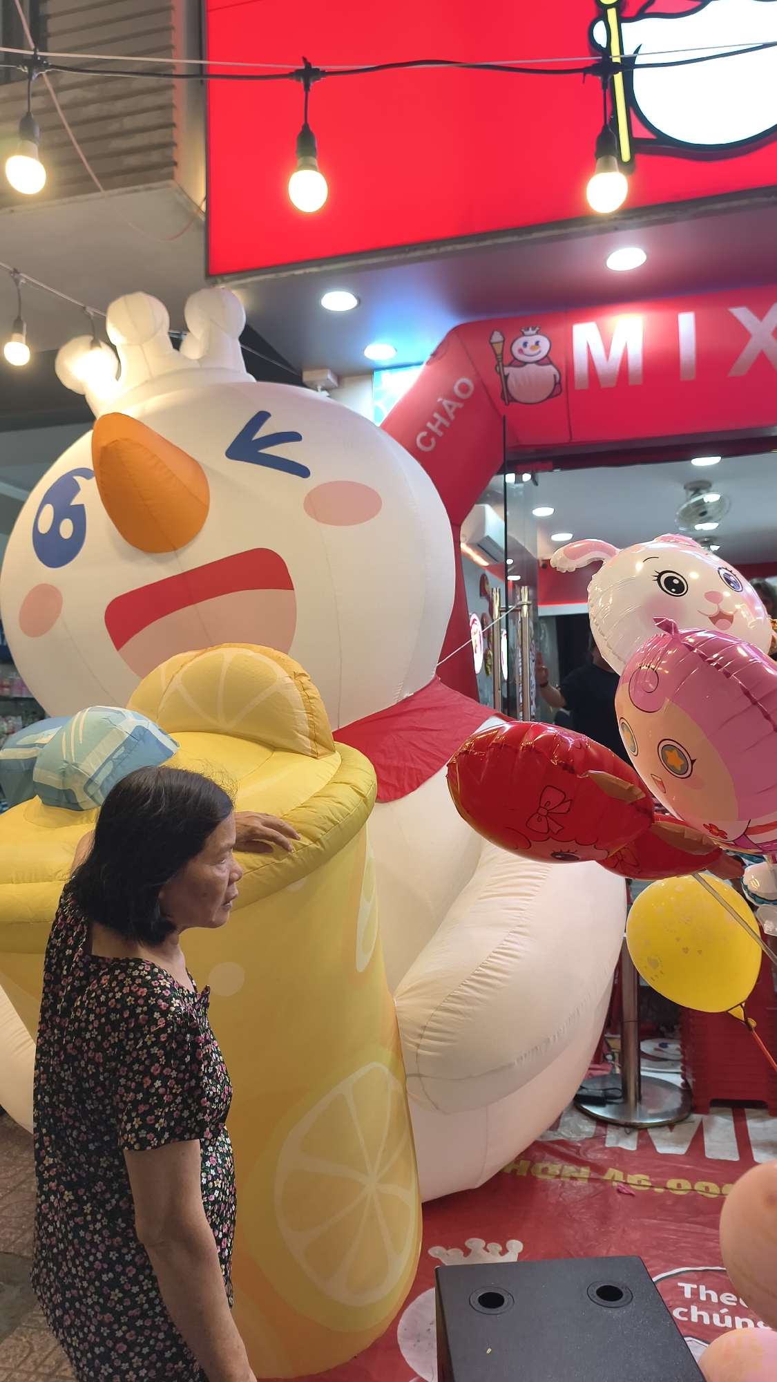 In an urban setting, a woman stands to the left of a large, inflatable character resembling a snowman. The snowman has a white body, a red scarf, and a large, smiling face with pink cheeks and blue eyes. To the right of the snowman, there are several balloons, including a pink pig and a white rabbit. Behind the snowman and balloons, an arched entranceway is visible with the word "MIX" in large, white letters on a red background. The overall scene appears to be outdoors, possibly in front of a shop or event space. The lighting suggests it is daytime.