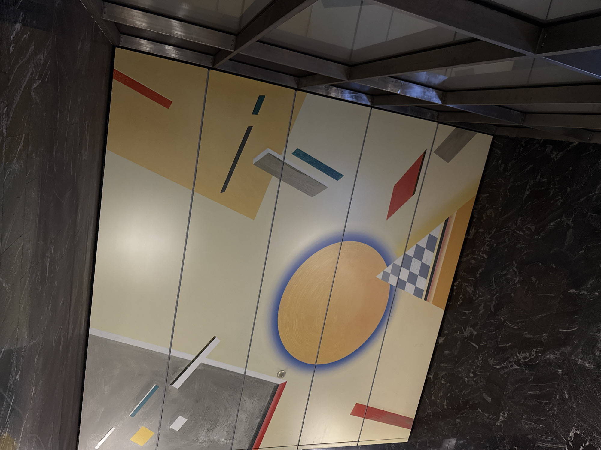 The image shows an abstract mural on a wall, possibly panels, under a modern ceiling structure. The mural has a light yellow-beige background with various geometric shapes in colors like red, gray, blue, yellow, and black/white checkerboard. A large, golden-yellow circle with a blue outline dominates the center. The mural is situated between dark marble-like walls.
The location is the Mayakovskaya metro station in Moscow, Russia.