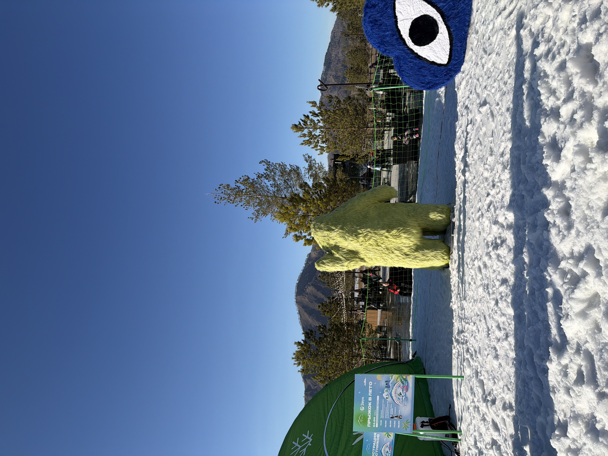 The scene depicts a snowy outdoor area in Озёрное, Russia under a clear blue sky. There are two large, furry character figures present - one blue with a prominent eye and another yellow. A green tent partially obscures the lower left, with a sign in Russian attached. Behind the figures and signage is a metal fence, trees, and blurred figures that appear to be people. A mountain can be seen in the background. The ground is covered in snow.