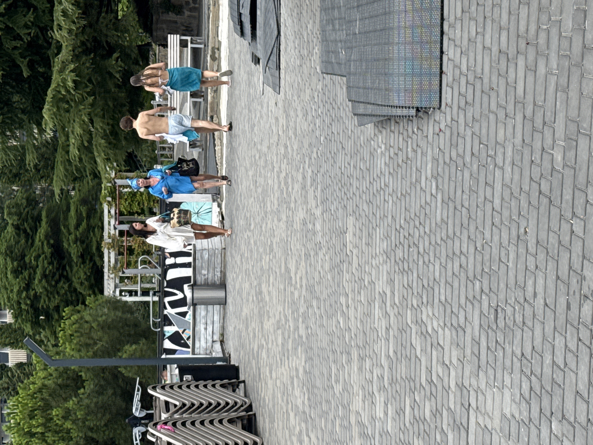 Four people are walking away from the camera on a paved walkway in an outdoor area. In the foreground, the walkway is paved with grey rectangular stones arranged in a diamond pattern. To the right of the walkway, several stacks of dark grey interlocking mats are present, appearing to be new or under construction.
The first person from the left is a woman wearing a white dress and carrying a large patterned bag. Next to her is another woman in a bright blue dress and a blue head wrap, also carrying a large black bag. Following them is a shirtless man wearing light blue shorts and a white towel around his waist. The fourth person is a woman wearing a white bikini top and a teal towel wrapped around her waist.
In the background, there are stacks of grey lounge chairs on the left. A metal trash can is visible between the chairs and the people. Behind the people, there is a low white wall with black and white abstract graphics on it. Further back, there are trees, a white bench, and some outdoor structures, possibly part of a resort or park. The overall environment appears to be a warm, sunny day. The location is described as Yalta, Ukraine.