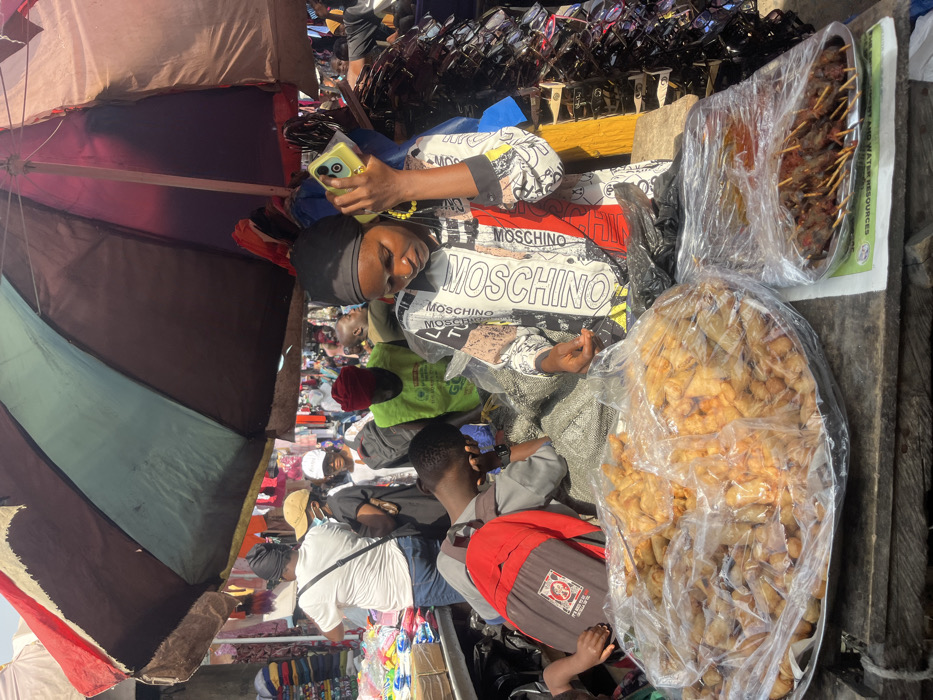 A busy outdoor market scene in Lekki, Nigeria, is depicted under the partial shade of a large, multi-paneled market umbrella featuring red, blue-green, and brown sections. In the foreground, a female vendor, wearing a black head covering and a "MOSCHINO" branded long-sleeved shirt, holds a yellow-cased smartphone to her left ear while looking downwards. Her right hand rests near a black plastic bag on a rough wooden surface. Directly in front of her, two silver platters, covered with clear plastic wrap, display food items for sale: one large platter contains numerous fried pastries (likely spring rolls), and a smaller platter holds several skewers of grilled meat. To the immediate left of the vendor, a person wearing a grey shirt and a red backpack leans over the wooden surface. The background reveals a dense market environment with multiple individuals, including another vendor in a white t-shirt sorting clothing to the far left, and a person in a green vest further back. Various stalls displaying merchandise, such as hanging clothing and racks of sunglasses, are visible, indicating a bustling commercial area. The scene is brightly lit by natural sunlight, casting distinct shadows.
