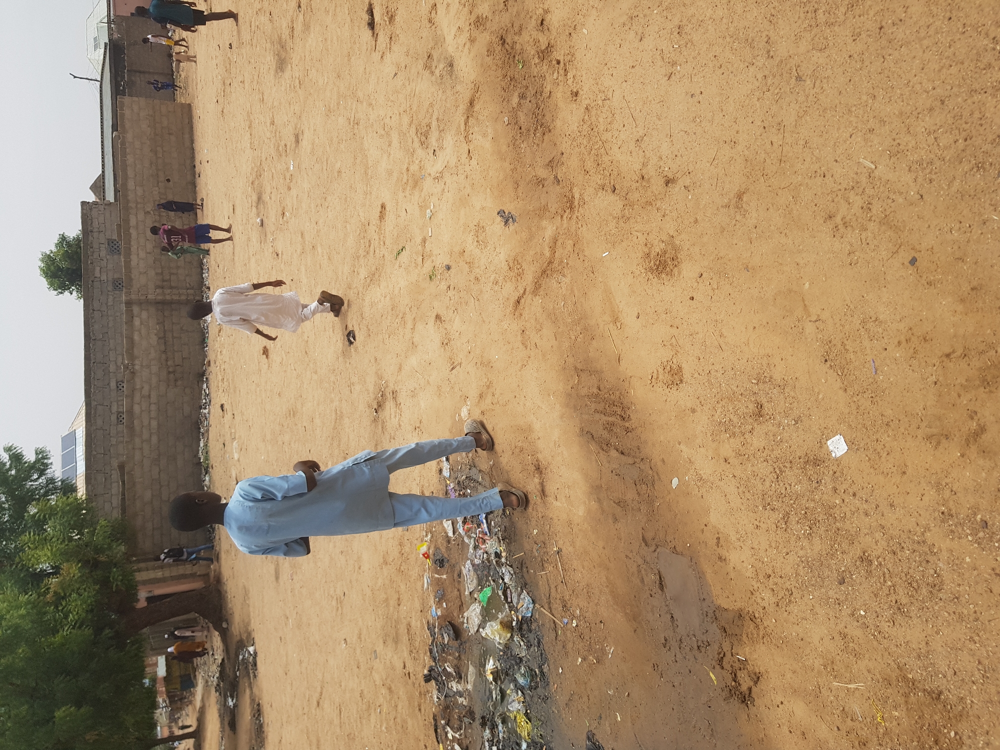 The image depicts an outdoor scene in what appears to be the city of Dutsinma B, Nigeria. The ground is primarily sandy with patches of dirt. A significant pile of garbage and stagnant water is visible in the lower left foreground.
Several individuals are present. A man in a light blue outfit stands near the garbage. Further in the background, another individual is wearing white, and what looks like children are playing a game in sportswear. A brick wall with a tree behind it is also visible in the background.