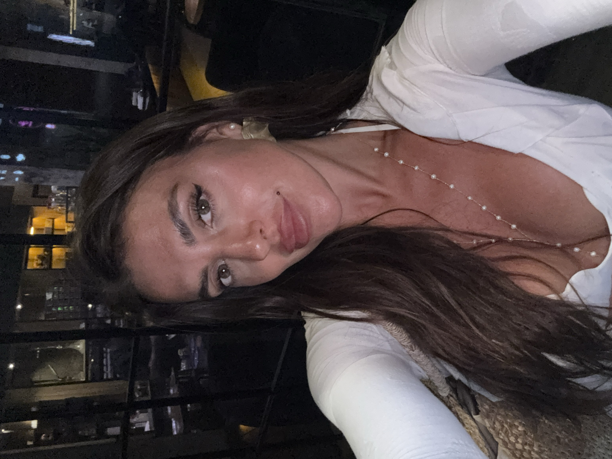 A woman with long, dark brown hair, wearing a white top and a pearl necklace, is taking a selfie in a dimly lit indoor setting, possibly a restaurant or bar. The background shows blurred interior elements with soft lighting and what appear to be shelves with bottles. The woman has brown eyes, winged eyeliner, and is wearing large, golden earrings. She is looking directly at the camera. The overall ambiance suggests a social evening setting.