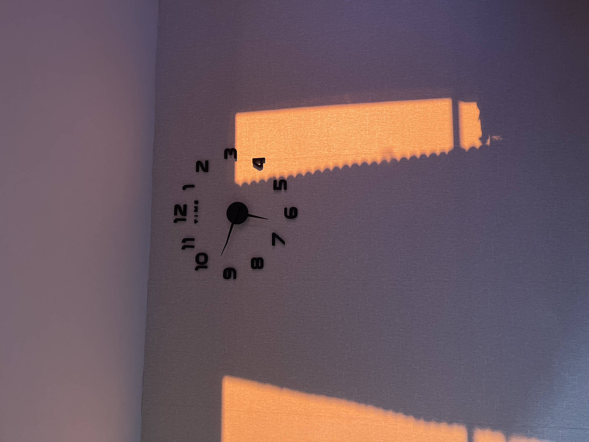 A wall clock with black numbers is on a wall with textured wallpaper. The clock reads approximately 6:36. Sunlight is cast onto the wall, creating a rectangular band of light with a serrated edge at the top and a less defined shape at the bottom. The light suggests an open window or blinds. The location is not identifiable based on the image alone.