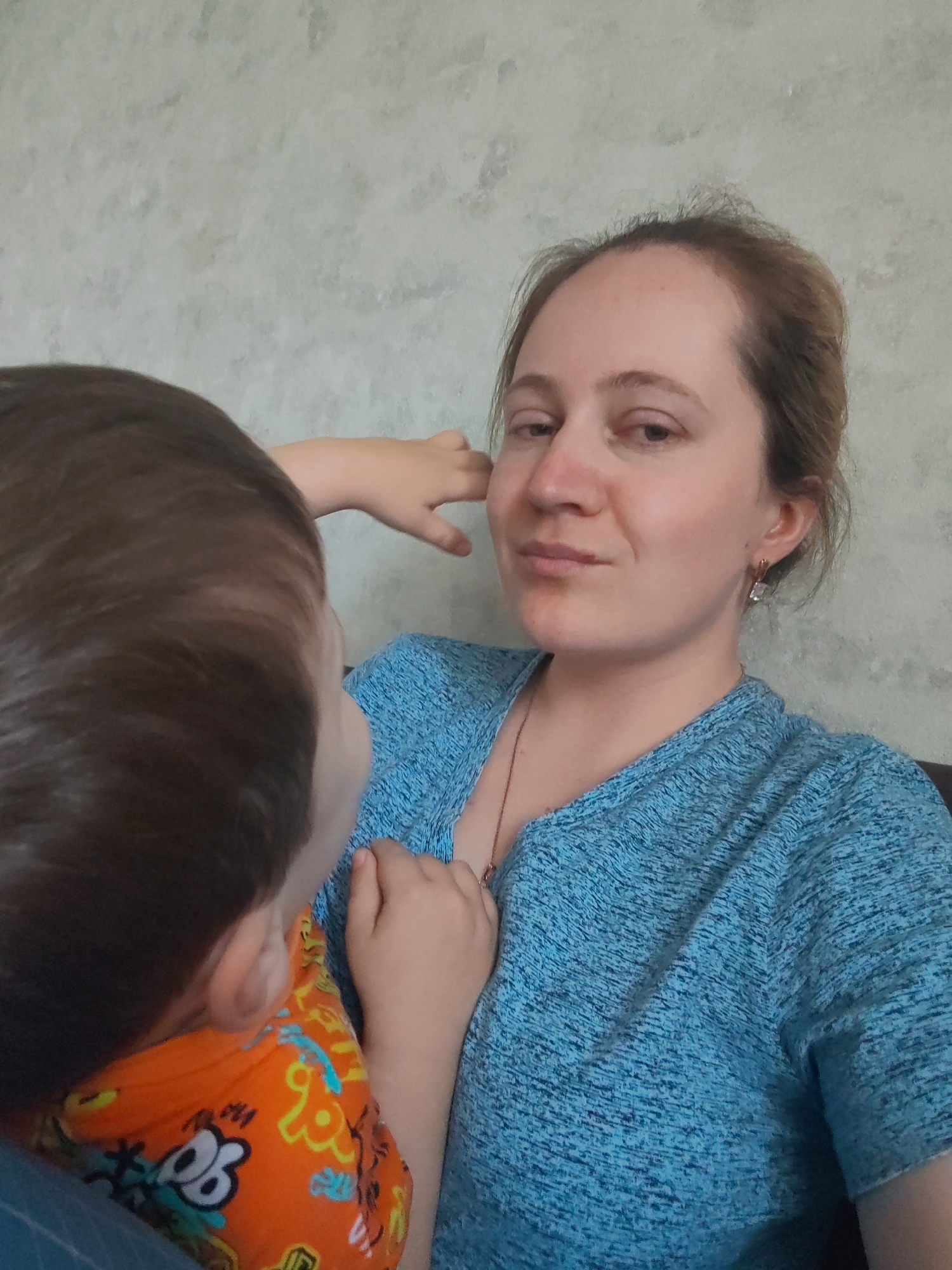The image shows a woman and a child. The woman is wearing a blue shirt and has her hand on her face. The child is wearing an orange shirt with cartoon characters. The background is a textured wall. The image appears to be a selfie taken in a home environment. It is not possible to determine the location in the city of Sokol, Russia, from the provided image.