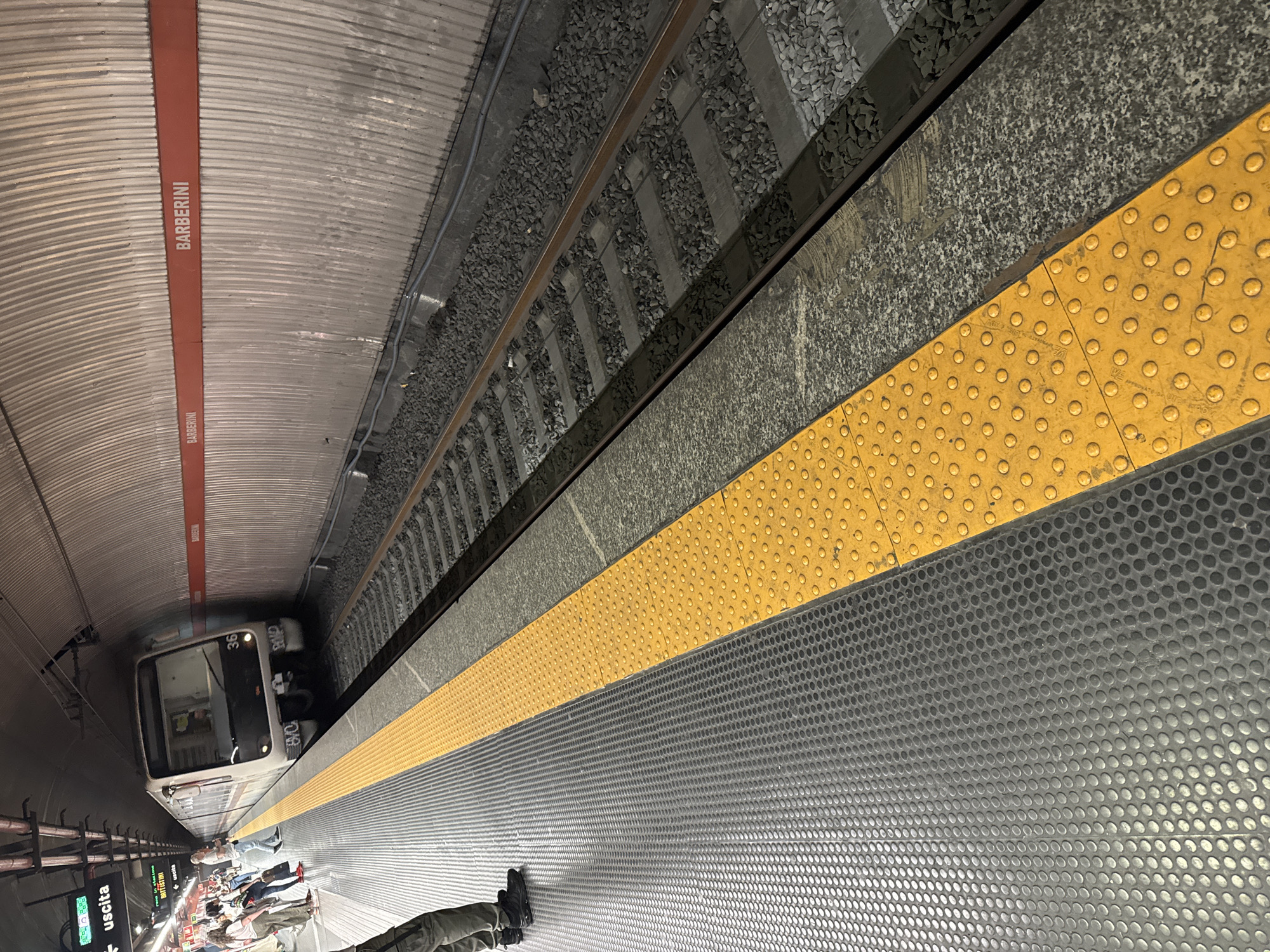 The image shows a subway platform at the Barberini station in Rome, Italy. The platform is gray with a textured surface and a yellow tactile strip along the edge. A subway train is approaching the platform. People are waiting on the platform. Above the platform, a sign reads "BARBERINI" in white letters on a red background. The subway tracks are visible next to the platform. The station's roof is curved and metallic.