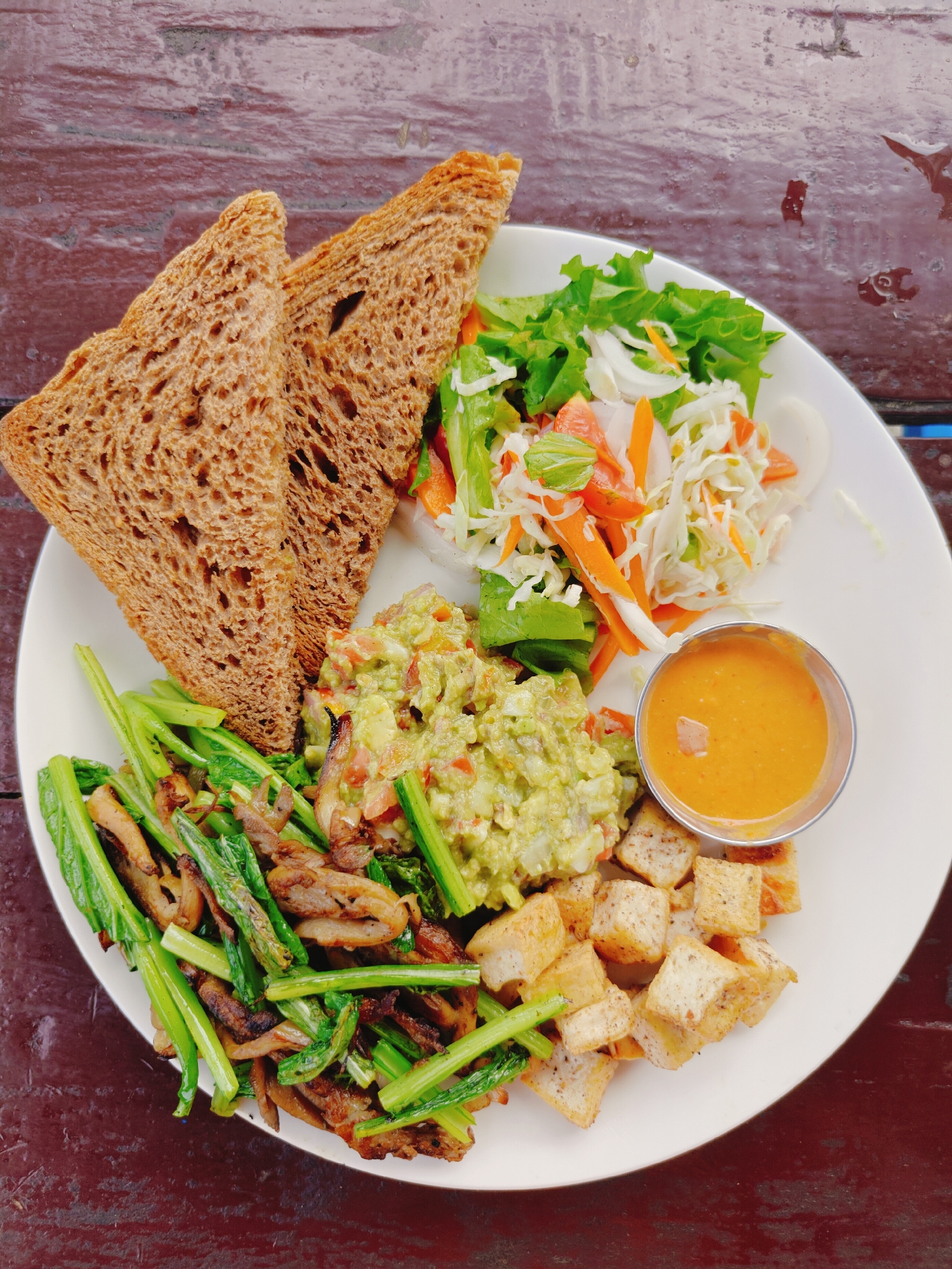 A white plate of food sits on a dark brown wooden table. The plate contains two slices of toasted whole wheat bread, a salad with lettuce, shredded carrots, and onions, guacamole with chopped tomatoes, a small metal cup of orange sauce, fried tofu cubes, and a mixture of stir-fried green vegetables and mushrooms. The table appears to be outdoors. The image is taken from a top-down perspective.