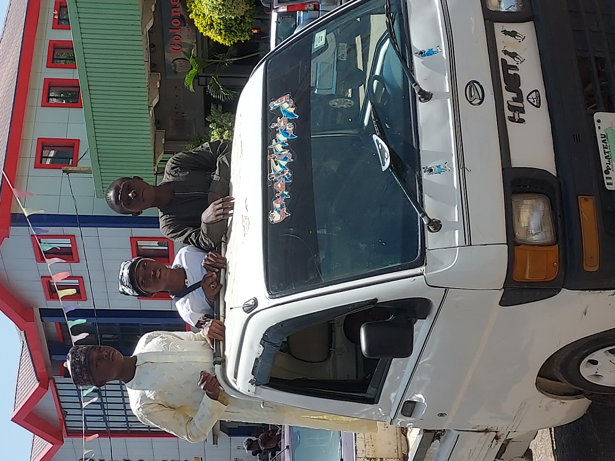 Three young men stand next to a white "HIJET" truck with a "PLATEAU" license plate. The truck has stickers on the windshield and hood. They are in an urban area, with a building featuring red-framed windows and decorative pennants visible in the background. The location is likely Jos, Nigeria, due to the PLATEAU license plate.
