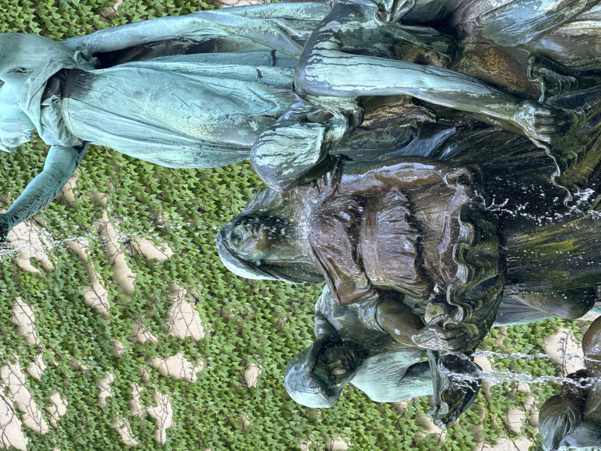 The image shows a detail of a bronze fountain sculpture situated against a wall covered in green ivy. The sculpture depicts several female figures, with water flowing over them and cascading down. One figure in the center appears to be holding a shell or basin, from which water is pouring. Another figure to the left is leaning forward, her face partially obscured. A larger figure stands behind and to the right, draped in flowing fabric, with her arm raised. The bronze has a weathered patina, with areas of green oxidation. The ivy on the wall is dense and lush, with individual leaves visible. The overall impression is one of movement and fluidity, enhanced by the presence of water. This sculpture is part of the fountain at the entrance to the Art Institute of Chicago, located at 111 South Michigan Avenue in Chicago, Illinois, United States of America.