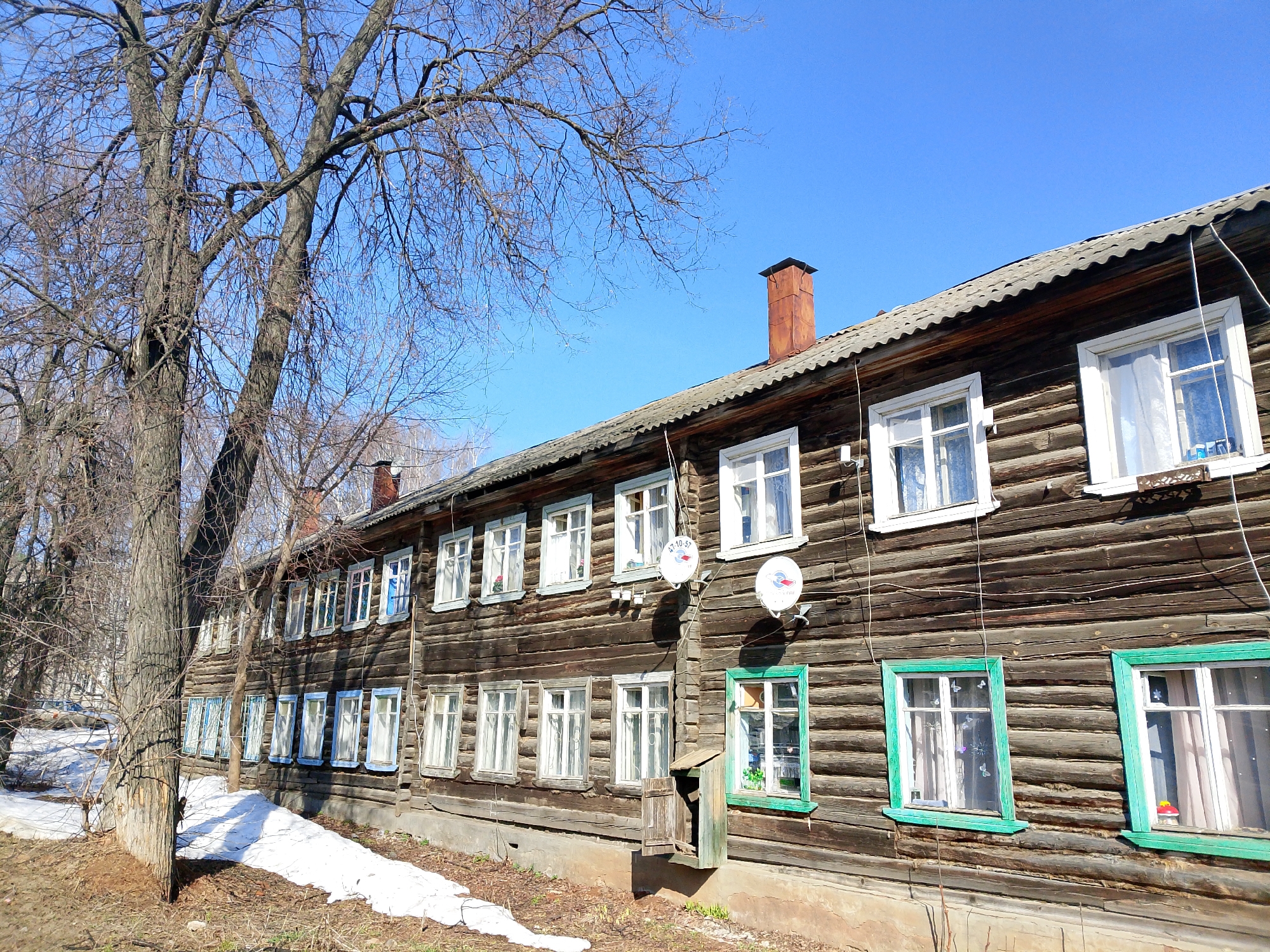 The image depicts a long, two-story, log-built residential building located in Izhevsk, Russia. The building is likely constructed from dark, aged wood. It has multiple windows with various colored frames, mostly white or blue, with a few painted green. Two satellite dishes are attached to the wall. A small, rustic mailbox or storage container is fixed below one of the windows. The roof is gray. A brick chimney protrudes from the roof near the center of the frame. In front of the building, there is a sparse landscape with a tree devoid of leaves and patches of melting snow on the ground, suggesting it is early spring. The sky is clear and blue. The building appears to be inhabited, evidenced by curtains and items visible in some of the windows.