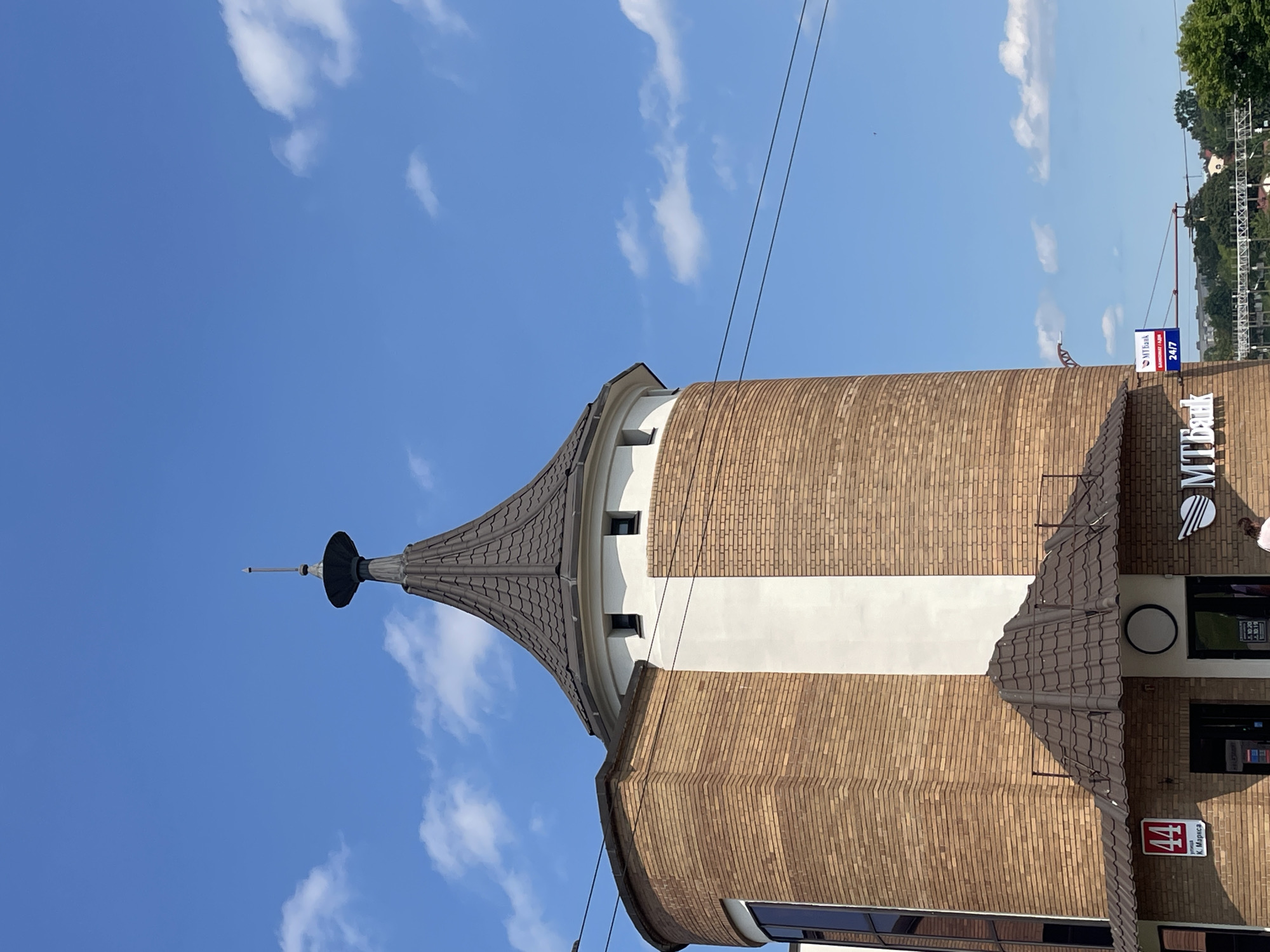 The image captures a close-up, eye-level view of a brick tower with a conical roof and a spire, set against a bright blue sky with scattered white clouds. The tower, situated in the city of Hrodna, Belarus, is constructed with light brown bricks and features a prominent white vertical stripe on its cylindrical section. Several dark, narrow windows are visible near the top of the tower, just below the roofline. A dark brown, tiled awning covers the entrance area at the base of the tower, which has dark doorways. A red sign with the number "44" and text below it is affixed to the brick wall above one of the entrances. To the right of the tower, a sign for "MTBank" is visible, along with a smaller blue and white sign indicating "24/7". Electrical wires are strung across the scene in front of the tower and the sky. In the background, a distant cityscape with trees and structures can be observed.