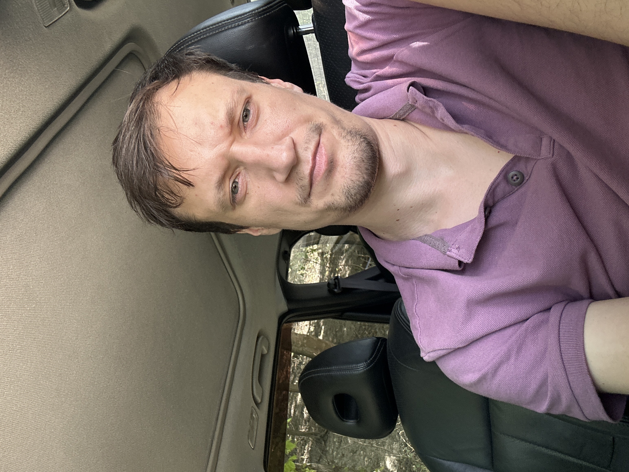 A medium shot shows a man with light skin and brown hair sitting in the driver's seat of a car. He is wearing a purple polo shirt. The man is looking at the camera with a slightly furrowed brow and a neutral expression. The car interior is visible, including the black leather seats and the sunroof. Outside the car, trees and foliage are visible, suggesting a rural or wooded area. The image appears to have been taken during daylight hours. The specific location in Makiv, Ukraine, cannot be determined from the visual information provided.