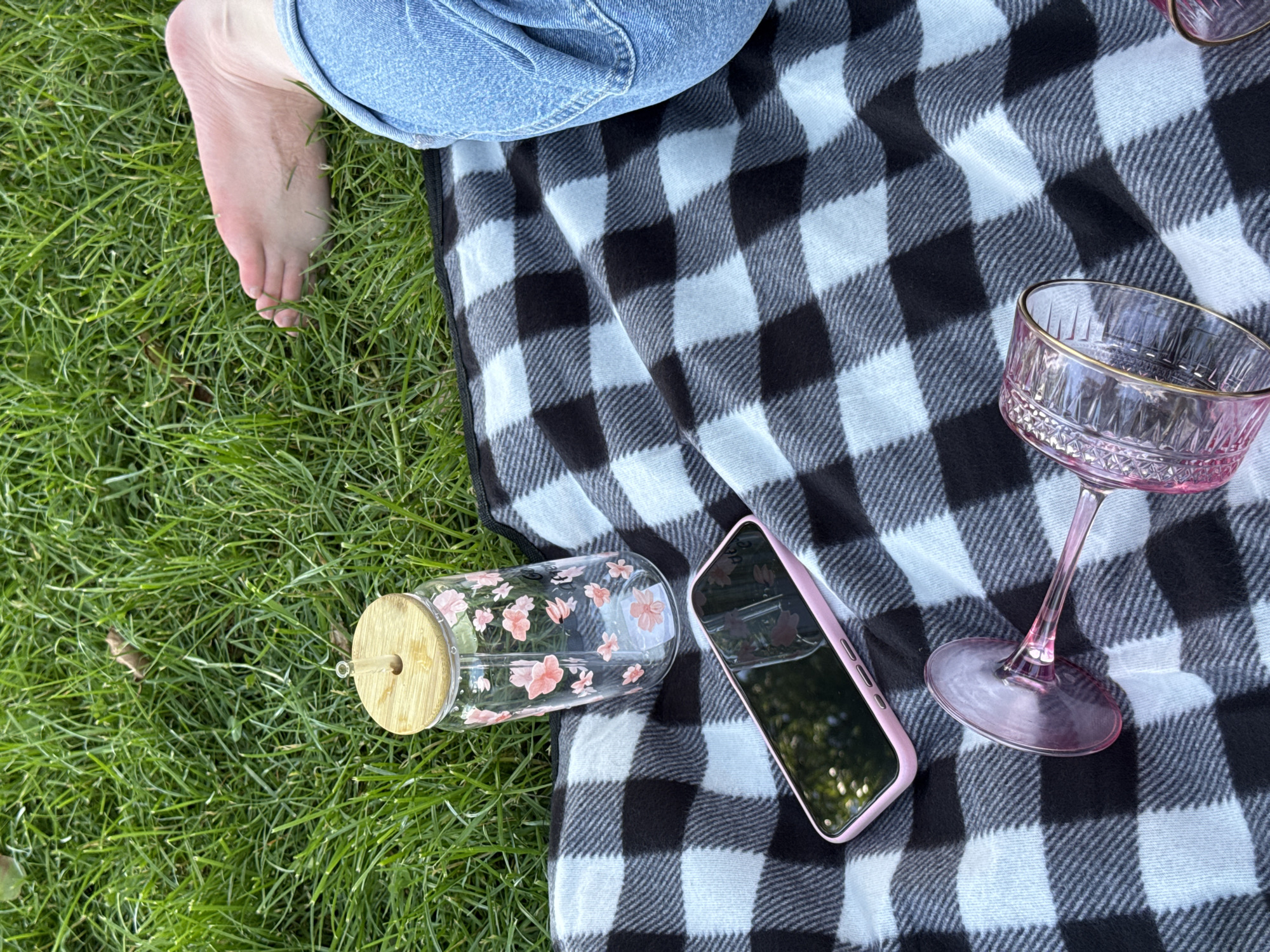An overhead, medium shot captures a picnic scene on a grassy lawn. A black and white checkered blanket is spread out, partially covered by lush green grass. On the blanket, a clear glass tumbler with a bamboo lid and a floral design sits upright. Beside it, a pink smartphone rests flat. In the foreground, two pink glass goblets with gold rims lie on their sides. To the right of the frame, a person's bare foot and lower legs, clad in blue denim jeans, are visible against the grass, suggesting they are reclining or lounging. The overall atmosphere is relaxed and leisurely.