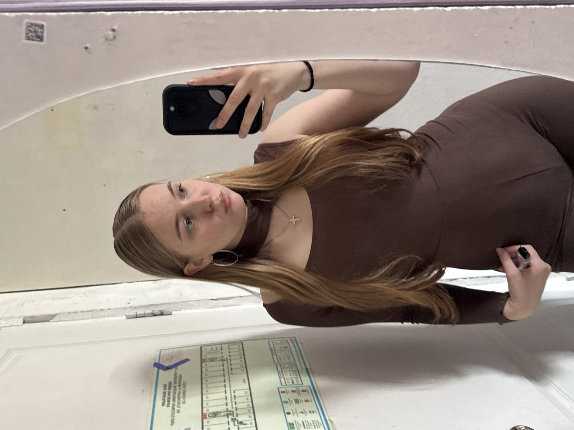 The image shows a young woman taking a mirror selfie. She is wearing a brown, long-sleeved, form-fitting jumpsuit. A thin, silver necklace with a cross pendant is visible around her neck, and she has a large hoop earring in her visible ear. Her long, blonde hair is parted in the middle and falls over her shoulders. She is holding a black smartphone in her right hand to capture the reflection. The background appears to be a bathroom or a similar indoor space, with a white, curved mirror and some textured white walls. Below the mirror, on a white surface that looks like a door, there is a sign with a diagram and text, possibly an emergency exit map or a safety instruction poster. Some of the text on the sign is visible, but not legible enough to be deciphered fully. The lighting suggests an indoor setting, likely during the day. The woman's expression is neutral.