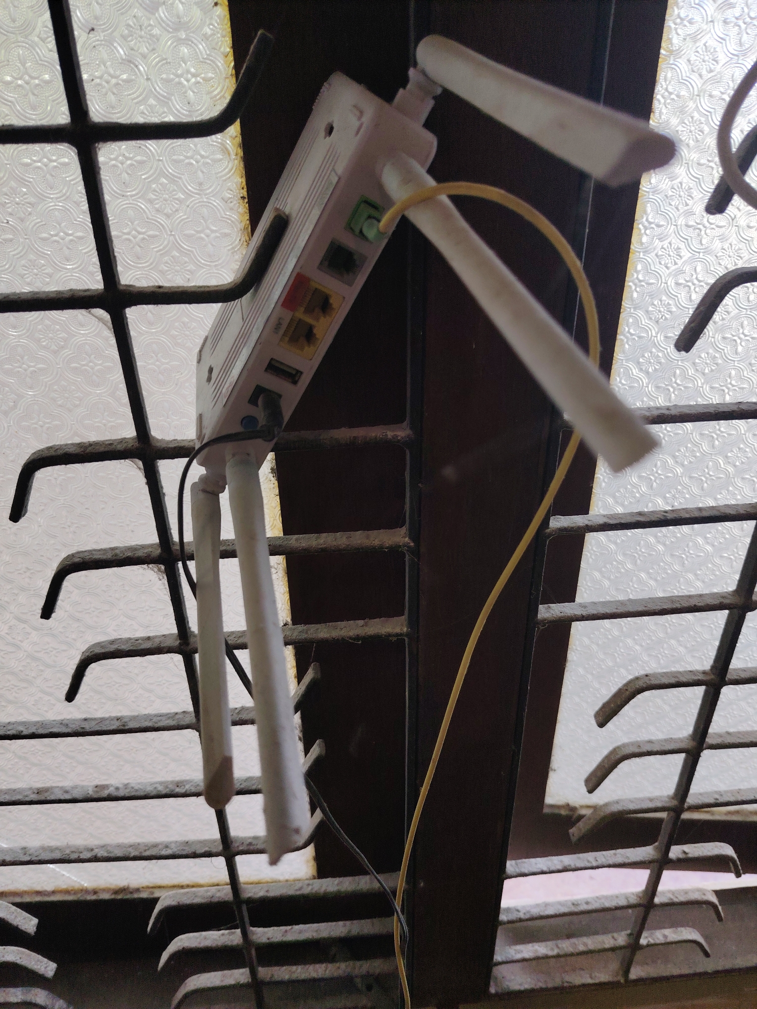 The image displays a white rectangular electronic device, likely a Wi-Fi router or extender, integrated into a dark, weathered metal window grill. The device features three white antennas, two angled upwards and one downwards.
On the side of the device, several ports are visible. These include a green Ethernet port, a yellow Ethernet port labeled "WAN", an orange Ethernet port labeled "LAN", a USB port labeled "USB", and a power input port labeled "DC IN". A yellow cable is plugged into one of the Ethernet ports, and a black power cable is connected to the DC IN port.
The window grill is made of dark, possibly rusty or dusty, metal, with horizontal and vertical bars; some horizontal bars have bent, hook-like ends. Behind the grill, a translucent glass pane with a repeating floral or geometric pattern diffuses light, indicating it is daytime. A dark brown frame or wall section is adjacent to the window. The setup suggests an indoor location, possibly an older building, where the device has been positioned in the window, likely for optimal signal.