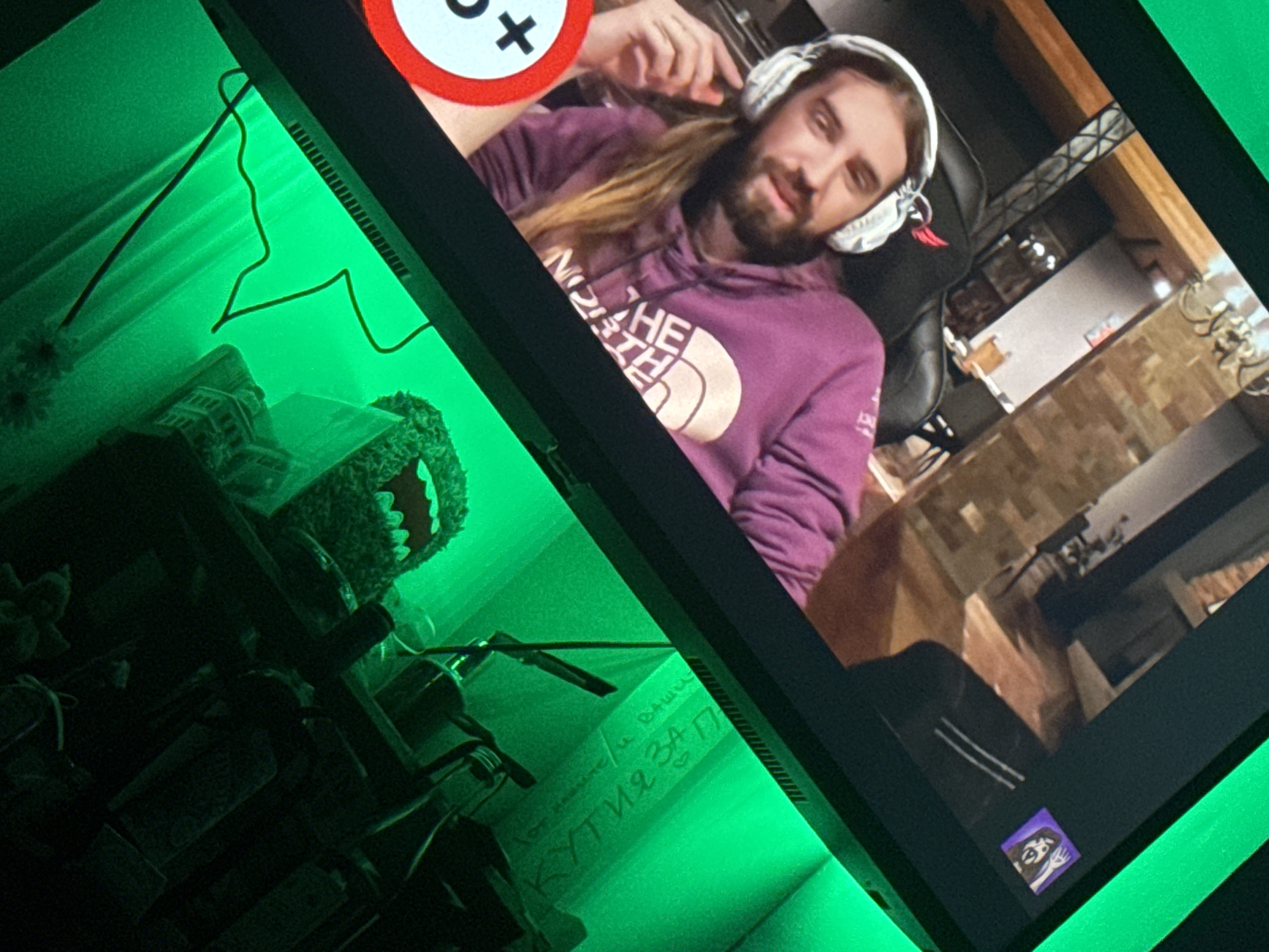 The image displays a person wearing headphones and a purple hoodie, visible on a computer monitor. The monitor is illuminated by green LED lights, creating a green glow on the left side of the frame. On the left side, a shelf is visible with various objects, including a plush toy and what appears to be a bundle of fabric or paper. There is also some handwritten text on a surface, partially illuminated by the green light. The text is in Cyrillic script and appears to be a quote. The monitor also has a small sticker with a drawing of a person on its bottom right corner. The overall scene suggests a home office or gaming setup. There is no discernible information about the location in Plovdiv, Bulgaria, nor any indication of the person's actions or interactions.