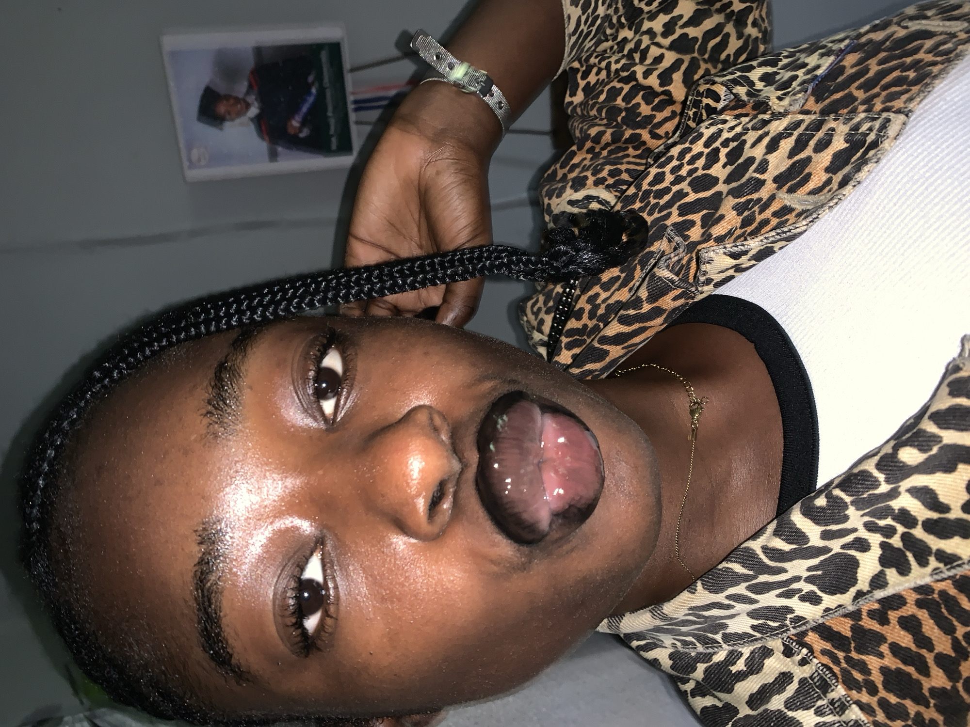 The image is a close-up selfie of a young Black woman with braided hair, wearing a leopard print jacket over a white top. She is wearing dark lipstick and has her tongue slightly out, appearing wet. A graduation photo is visible in the background on the wall, which appears to be in a residential setting. The lighting is dim and appears to be from an artificial source. The overall scene suggests an informal indoor setting, likely a home environment. No specific details about Rafin Par, Nigeria are discernible from the image itself.