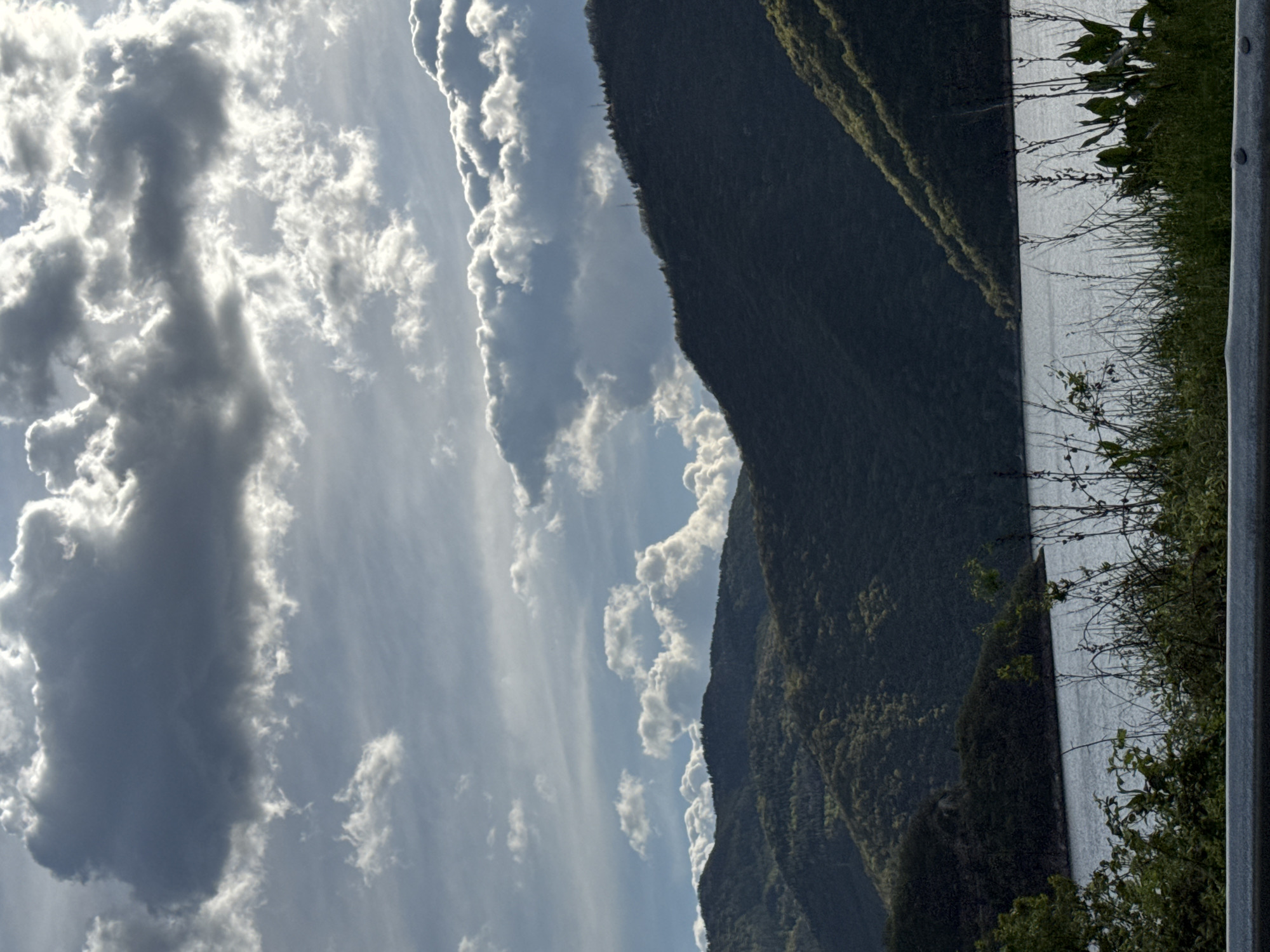 The image shows a mountainous landscape beside a body of water. The mountain is covered in dense vegetation. The water is calm, reflecting light. To the side, there's green vegetation with a gray post, possibly a roadside barrier. The sky is filled with clouds. The image could be taken near Mihalkovo, Bulgaria, due to the mountainous terrain and presence of water bodies.