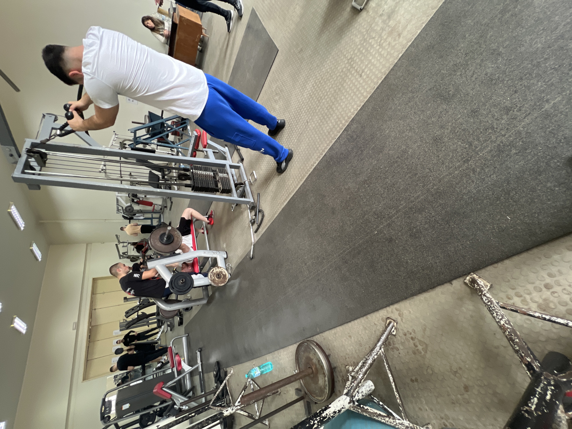 **Scene Description:**
**Location:** Gym in Sofia, Bulgaria.
**Environment:** The gym has gray rubber matting for flooring and a light yellow wall. There are fluorescent lights visible on the ceiling.
**People:**
* A man in a white t-shirt and blue sweatpants is performing a cable row exercise on a weight machine.
* Another man in a black t-shirt is using a leg press machine.
* A man in a white shirt is using a chest press machine.
* Other individuals are visible in the background, including a woman standing near a desk and other gym-goers near various exercise machines.
**Objects:**
* Various weight machines, including cable row, leg press, and chest press.
* Weight plates.
* Barbell on rack.
* A water bottle.
**Actions:**
* People are engaged in various weightlifting exercises.
**Interactions:**
* People are using the gym equipment to perform their workouts.