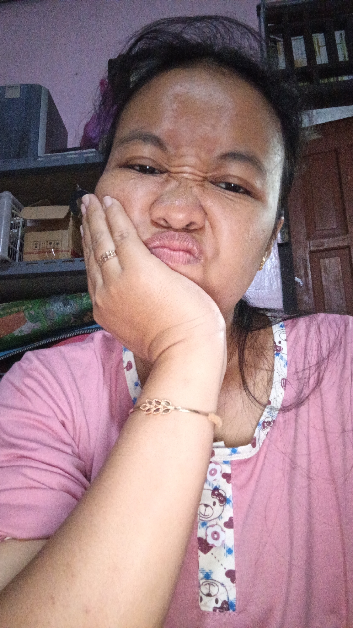 A young woman is taking a selfie indoors, her left hand supporting her face as she makes a pouting expression. She is wearing a pink t-shirt with a white collar adorned with cartoon bears and hearts. A gold-toned bracelet with a leaf design is visible on her left wrist, and a matching ring is on her left ring finger. The background shows shelves with various items, including boxes, and a wooden door. The overall setting appears to be a home environment.