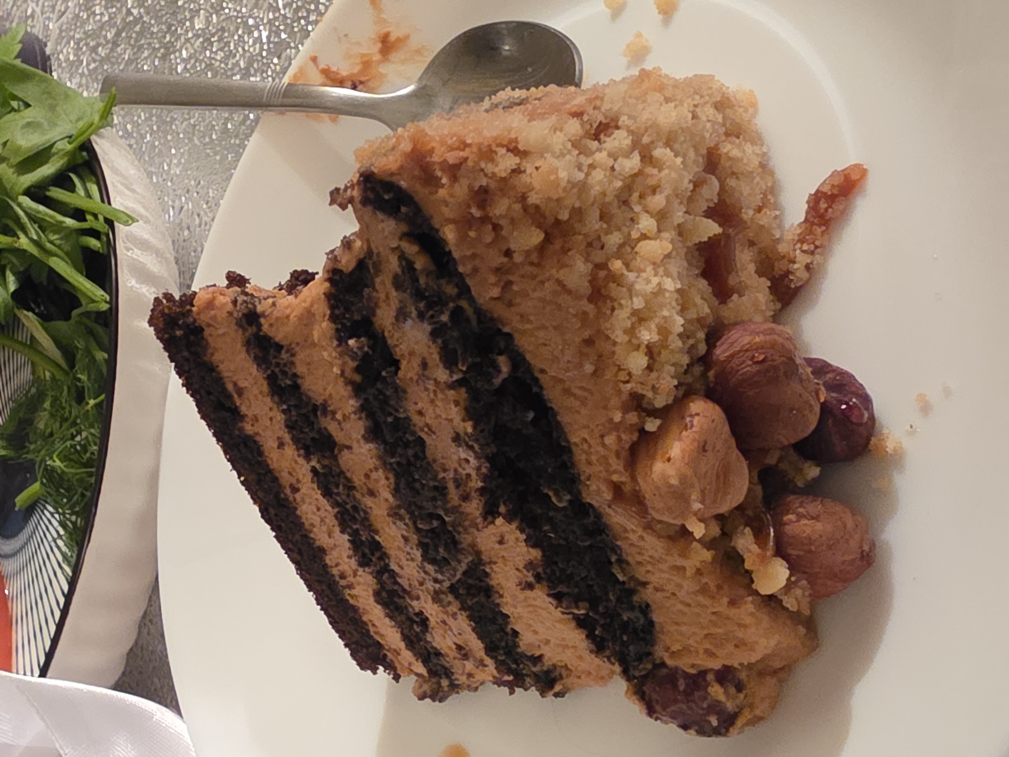 A slice of layered chocolate cake with tan frosting sits on a white plate. The cake has multiple layers of dark chocolate cake and lighter tan frosting. The outer edges of the slice are coated in a crumbly, tan mixture and whole hazelnuts. A spoon is visible to the right of the cake. In the background, a white bowl contains green leafy vegetables, and a white ribbon is draped over the left edge of the frame. The setting appears to be indoors, possibly in a dining area in the city of Nagorny, Russia.