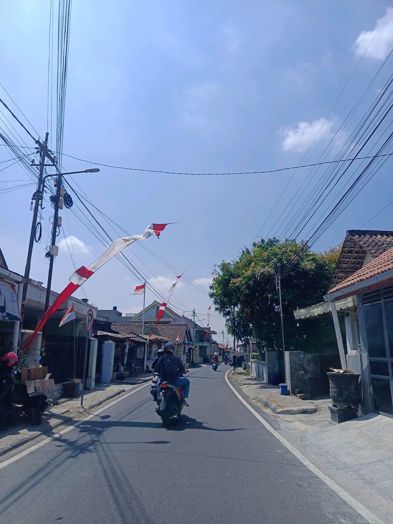 The image depicts a street scene in Tambran, Indonesia, under a clear blue sky with scattered white clouds. Overhead, a dense network of electrical wires is strung between utility poles. Red and white Indonesian flags are prominently displayed, draped along the street and tied to poles, suggesting a national holiday or celebration.
The street itself is paved with asphalt and has a white line marking the edge of the lane. On the left side of the road, there are buildings with various architectural styles, some appearing to be houses or small shops, with fences and parked motorcycles. One building features posters on its exterior. On the right side of the road, there are more buildings with tiled roofs and a large tree with green foliage.
Two individuals are riding motorcycles in the same direction down the street. The rider in the foreground is wearing a dark shirt, blue jeans, and a helmet, with a backpack visible. The motorcycle they are riding is dark in color. Another motorcycle is further ahead. The scene appears to be a typical day in a residential or commercial area of Tambran.