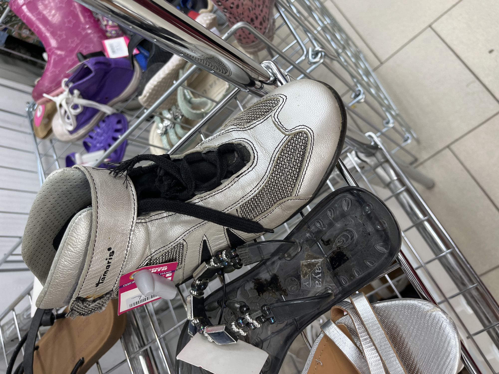 The image depicts a retail display of various footwear items on a multi-tiered metal wire shelving unit within a store located in the city of Minsk, Belarus. The foreground is dominated by a silver metallic Tamaris brand high-top shoe, exhibiting textured side panels and a smoother toe cap, secured with black laces and bearing a white security tag with a visible "SECOND HAND" label. Adjacent to it, a black transparent "aya" brand jelly sandal with beaded and metallic strap embellishments is positioned beneath, showing visible wear and debris on its sole. A portion of a silver textured flat sandal with a tan insole is also visible in the lower-left corner. In the background, multiple blurred footwear items are discernible on the shelves, including a brown flat sandal, a bright purple low-top sneaker with white laces, a purple plastic sandal, and a pink rain boot adorned with star patterns. The shelving unit is constructed from chrome-colored metal wires, and the floor beneath consists of light-colored tiles. No individuals are visible in the frame.