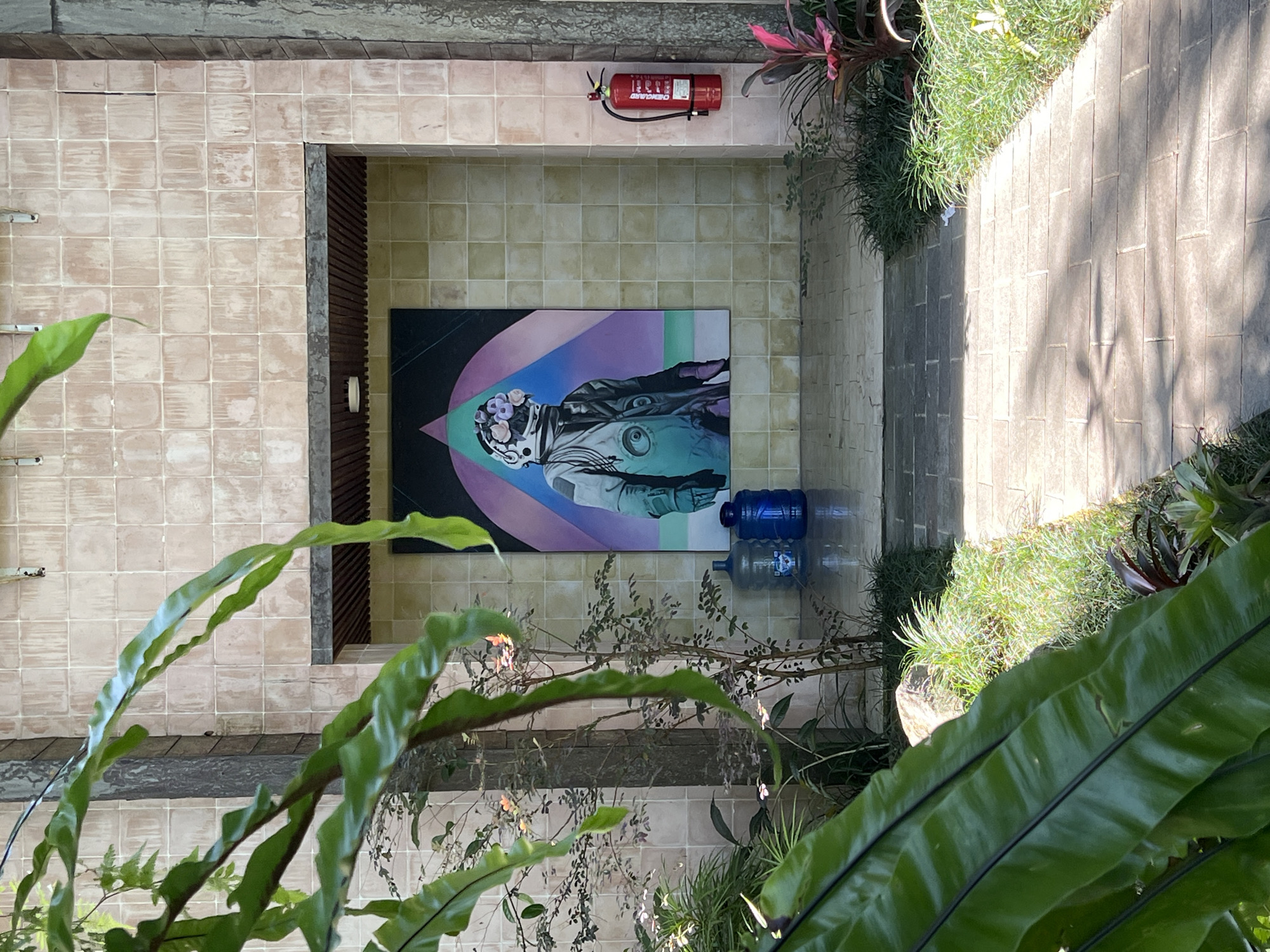 The image, seemingly taken from above in Petulu, Indonesia, reveals an interior courtyard space composed of tiled flooring. A painting is displayed on one of the walls, depicting an astronaut figure. Above the painting is a red fire extinguisher. The scene is framed by lush greenery. In the lower right are large green leaves. To the right is concrete and plants. In the lower center are a collection of thin leaved bushes and plants. Two blue water jugs sit on the floor in front of the painting.