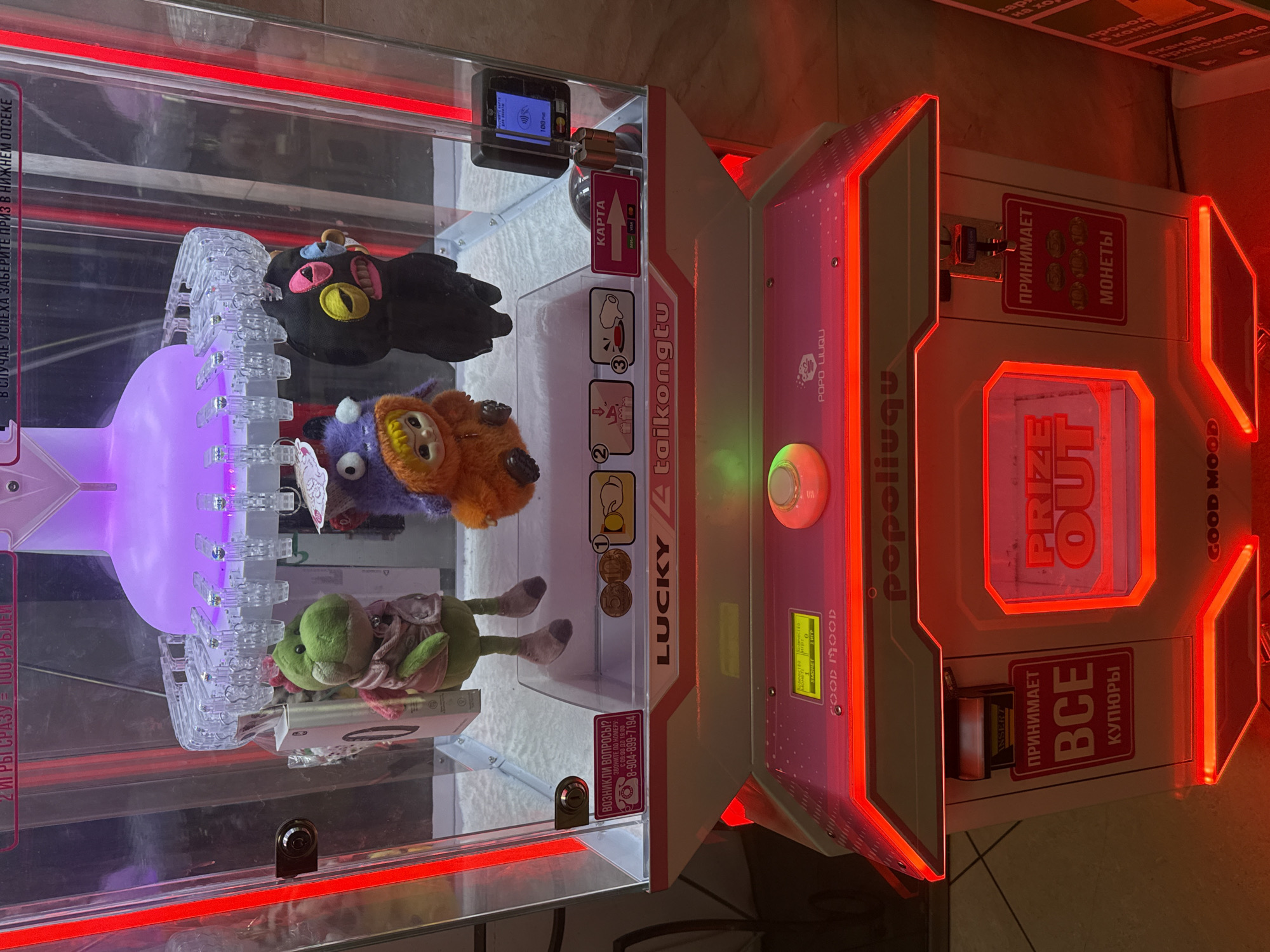 The image shows a claw machine arcade game with stuffed animals inside. The machine is brightly lit with red and purple lights. The game has a touchscreen display and a coin slot. There are three stuffed animals visible: a green dinosaur, a purple creature with an orange face, and a black creature with a pink mouth and yellow eyes. The machine is located in Norilsk, Russia. The overall environment appears to be an indoor public space, possibly a shopping mall or entertainment center, with a tiled floor visible in the background.