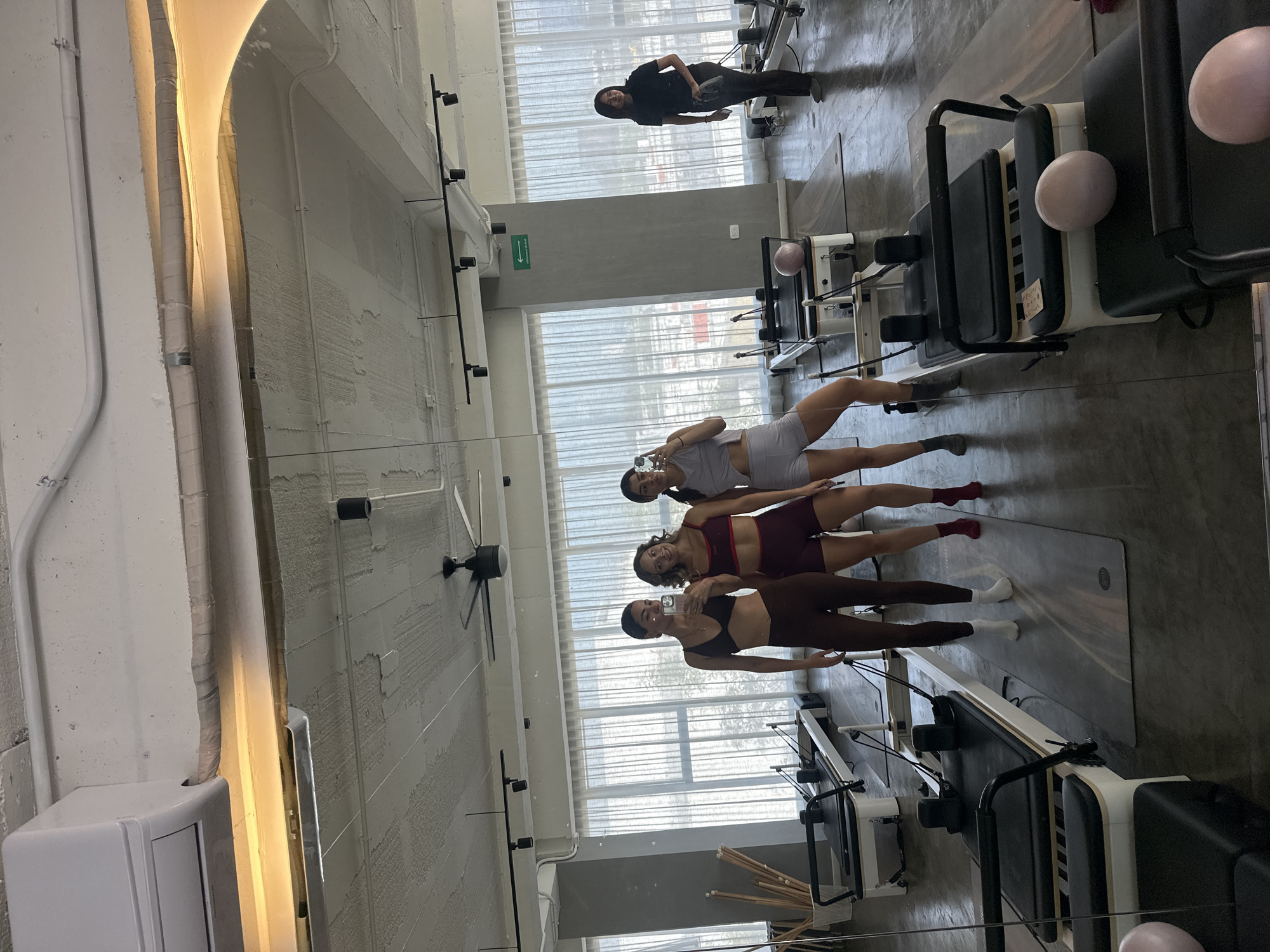 In a pilates studio in Puebla, Mexico, four women are participating in a class. The studio has a modern, industrial aesthetic with concrete walls and floors, and large windows draped with sheer white curtains. The room is equipped with multiple pilates reformers, some of which are visible in the foreground and reflected in the large mirror that lines one wall.
Three women are clearly visible in the reflection, dressed in athletic wear. One woman in the foreground is wearing a brown sports bra and matching leggings. Next to her is a woman in a burgundy sports bra and matching shorts, and another woman in a light purple sports bra and shorts. They appear to be in the middle of an exercise, with their bodies positioned on pilates mats. The fourth woman is less visible, seen from behind wearing a black hoodie and pants, standing near a reformer in the background.
The lighting suggests it is daytime, with natural light streaming through the windows. The atmosphere appears focused and professional, as expected in a fitness class. There are no obvious emotional cues beyond concentration. A pink pilates ball is visible on one of the reformers. A green directional arrow sign is mounted on the wall.