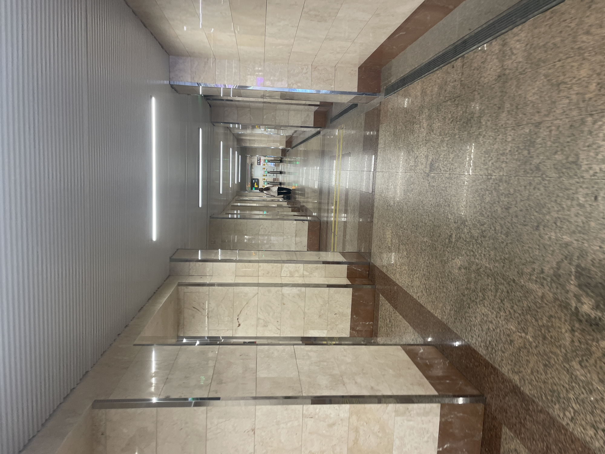 The image depicts a long, brightly lit corridor within a building in Sochi, Russia. The walls are tiled with light-colored stone, interspersed with mirrored panels and linear lighting. The floor is tiled in a darker, speckled pattern, with brown accents along the edges. A few people are visible in the distance, appearing as small figures at the far end of the corridor. The perspective emphasizes the length and symmetry of the hallway.