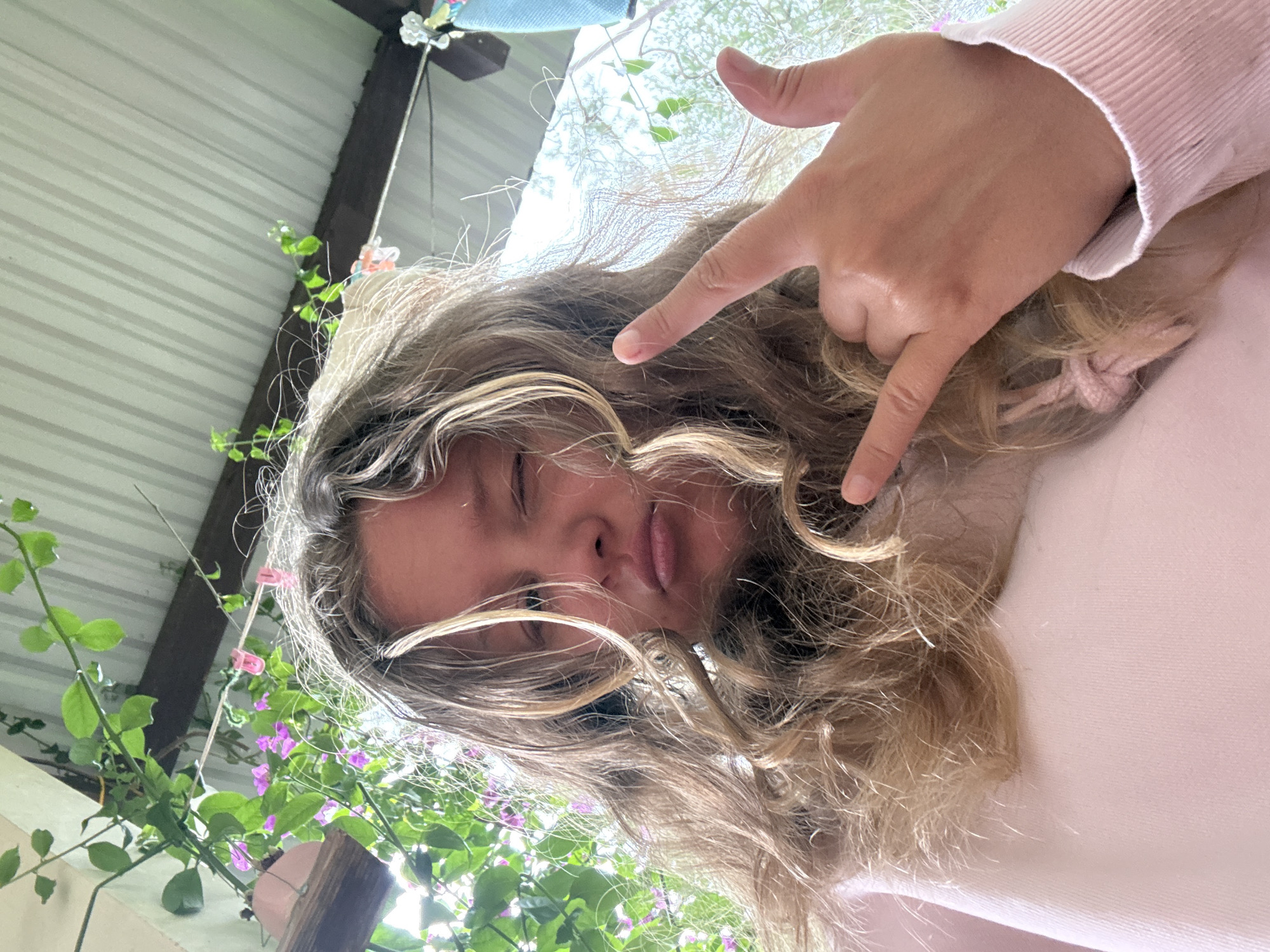 A young person with long, curly blonde hair and a pink sweatshirt is taking a selfie outdoors. They are winking and making a rock-and-roll hand gesture. The background features green vines with purple flowers, a corrugated metal roof, and wooden beams. The scene appears to be a residential outdoor area.