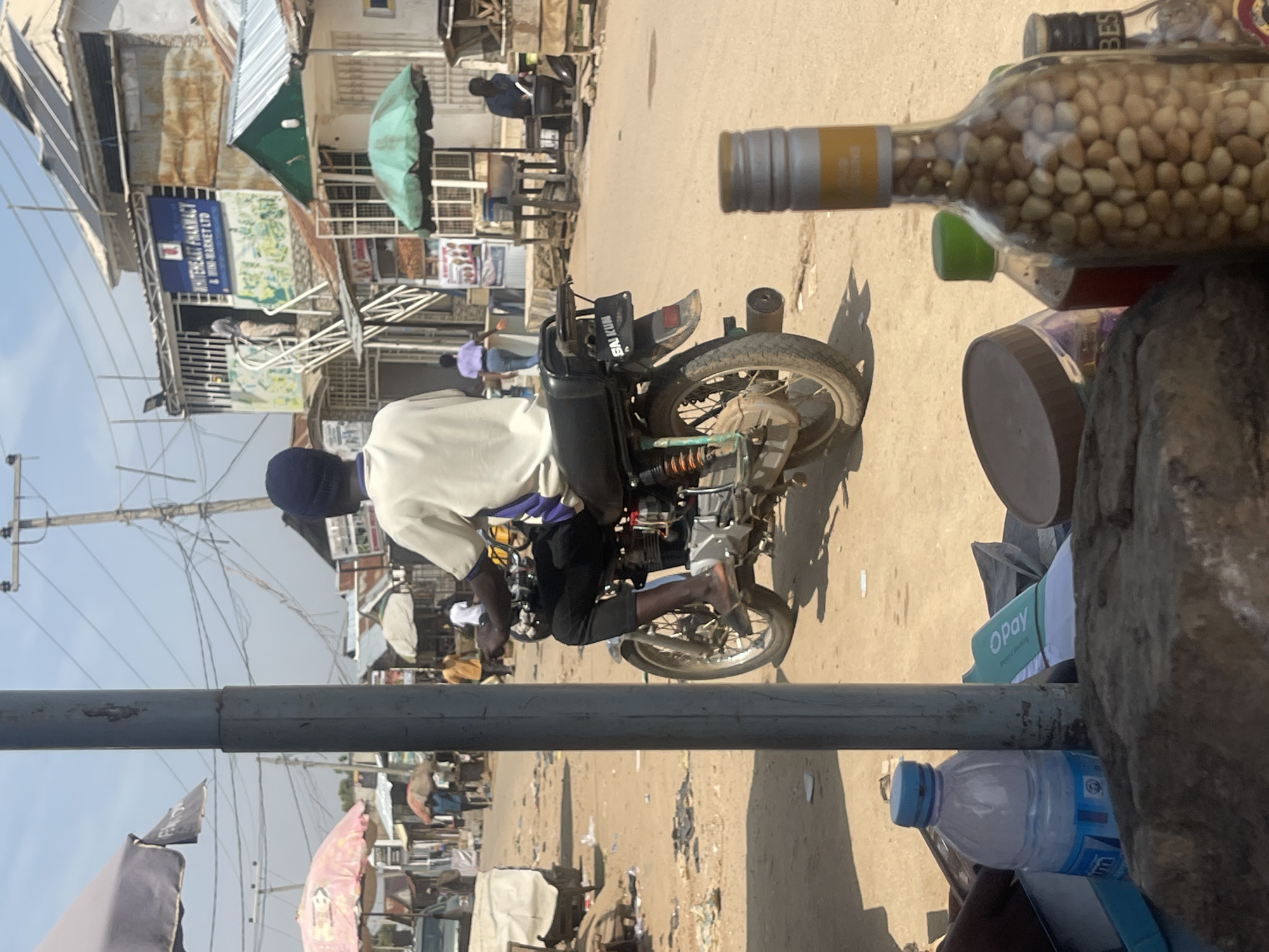 The image depicts a street scene in Abuja, Nigeria. In the foreground, a man wearing a dark blue head covering, a cream-colored shirt with purple trim, and black pants rides a motorcycle. The motorcycle is black and has a license plate with the inscription "SAI KUM". The road is unpaved and dusty, with scattered debris.
On the left side of the image, a gray metal pole stands prominently. In the background, several market stalls are visible. One stall features a blue sign that reads "WHITEHEART PHARMACY & MINI-MARKET LTD". Other stalls have various goods displayed under umbrellas. People are seen walking and interacting within the market environment.
To the right of the motorcycle rider, a green umbrella shades a stall where a person is seated. In the very foreground, a glass bottle filled with peanuts is visible, along with other items including a plastic water bottle and a green object with the inscription "OPay". The overall impression is a bustling, typical street market scene.