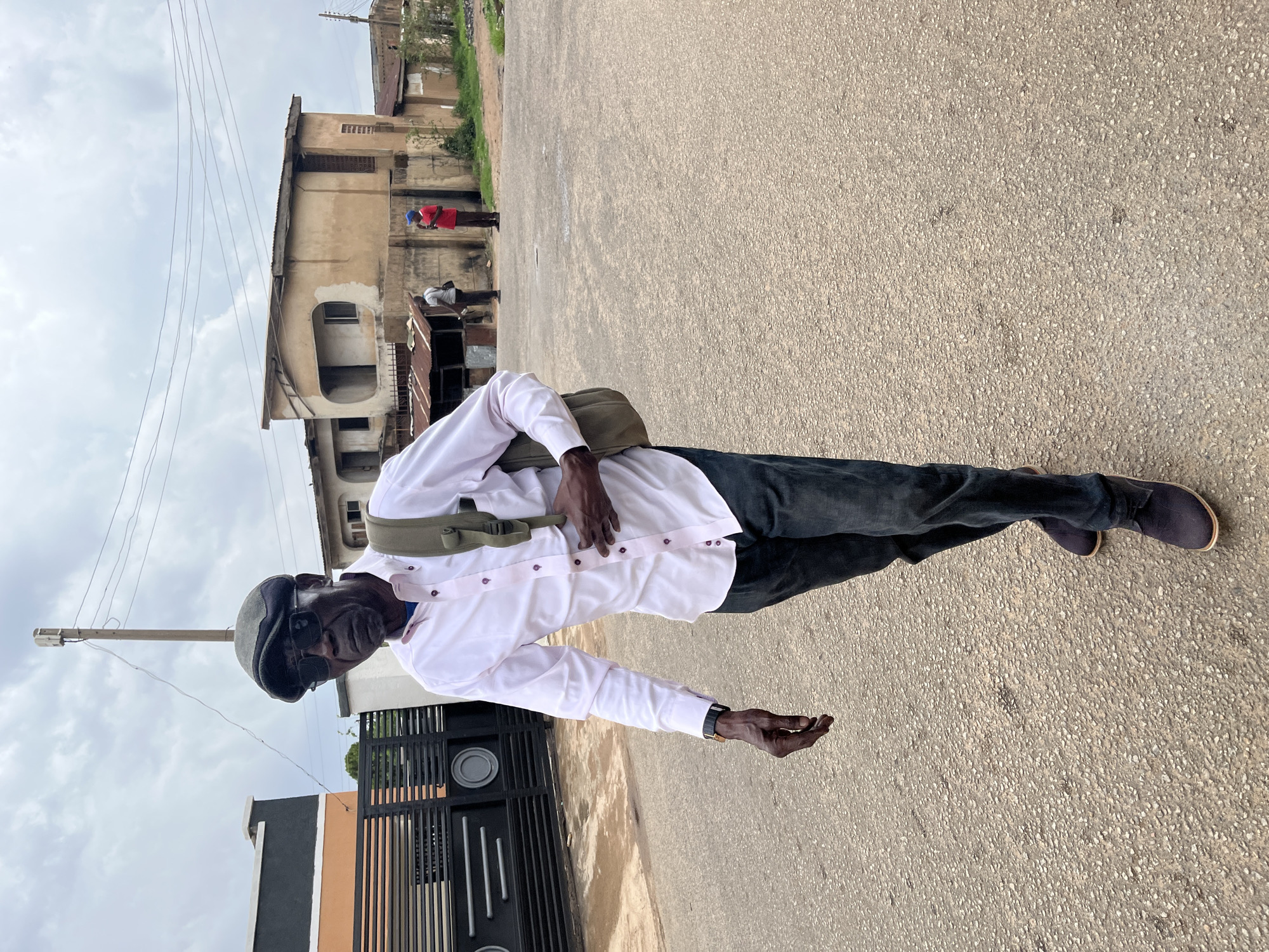 A portrait-style photograph captures a Black man standing on a paved street under an overcast sky in Jos, Nigeria. The man, appearing middle-aged to older, is dressed in a white long-sleeved button-down shirt with light purple buttons, dark trousers, and dark shoes. He wears a dark grey flat cap and sunglasses perched on his head. An olive green crossbody bag is slung over his right shoulder. His right hand rests near his midsection, while his left hand is held slightly open, palm facing upwards, with a dark watch visible on his wrist. He gazes directly at the camera with a neutral to slight smiling expression.
The street surface is light grey asphalt with small embedded stones. In the background to his left, there's a modern black metal gate, partially obscuring a building with orange and dark grey sections. Further down the street, on the opposite side, older multi-story buildings are visible, characterized by their light brown stucco-like facades and arched window openings. Several people are discernible in the distance near these buildings, including one person in a red shirt and blue cap, and another in a white shirt behind what appears to be a small market stall or structure. The sky is filled with light grey clouds, suggesting an overcast day. Numerous thin utility wires crisscross the upper portion of the frame, and a wooden utility pole stands behind the man's head. No visible text is present in the image.