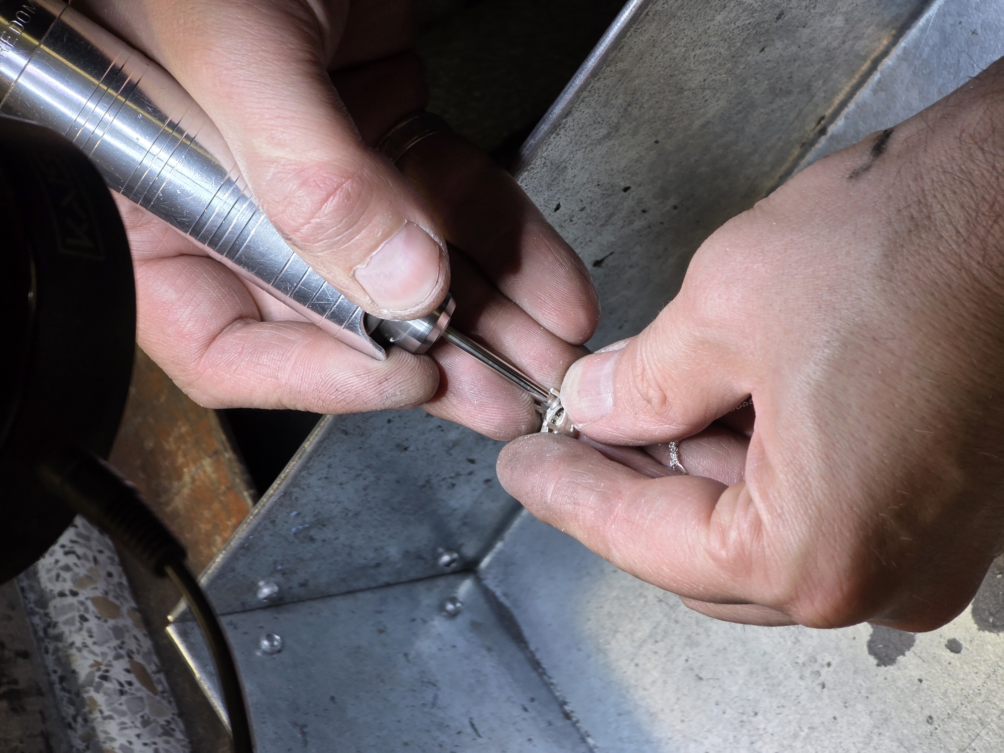 This is a close-up, high-angle shot of a person's hands working on a small, intricate object, likely jewelry. The subject is in the process of engraving or polishing a silver ring. One hand holds a rotary tool with a fine tip, while the other hand delicately manipulates the ring. The tools and the metal object suggest a workshop or a crafting environment.
The setting appears to be indoors, with a rough, metallic surface forming a corner of a structure, possibly a workbench or a specialized tool. There are hints of a textured floor and a darker, possibly wooden, surface in the background. The lighting is bright, highlighting the metallic sheen of the tools and the ring.
The focus is on the meticulous detail of the work being done, emphasizing the skill and precision involved. The visible skin texture on the hands, including fingerprints and slight abrasions, adds a sense of realism to the scene. The overall mood is one of focused concentration and skilled craftsmanship. There is no clear indication of time of day or weather.
While there is some text on the rotary tool, it is not clearly legible in the provided image. The location context of Kazan, Russia, does not offer any specific visual cues within the image itself, as the scene is generic to many crafting environments.