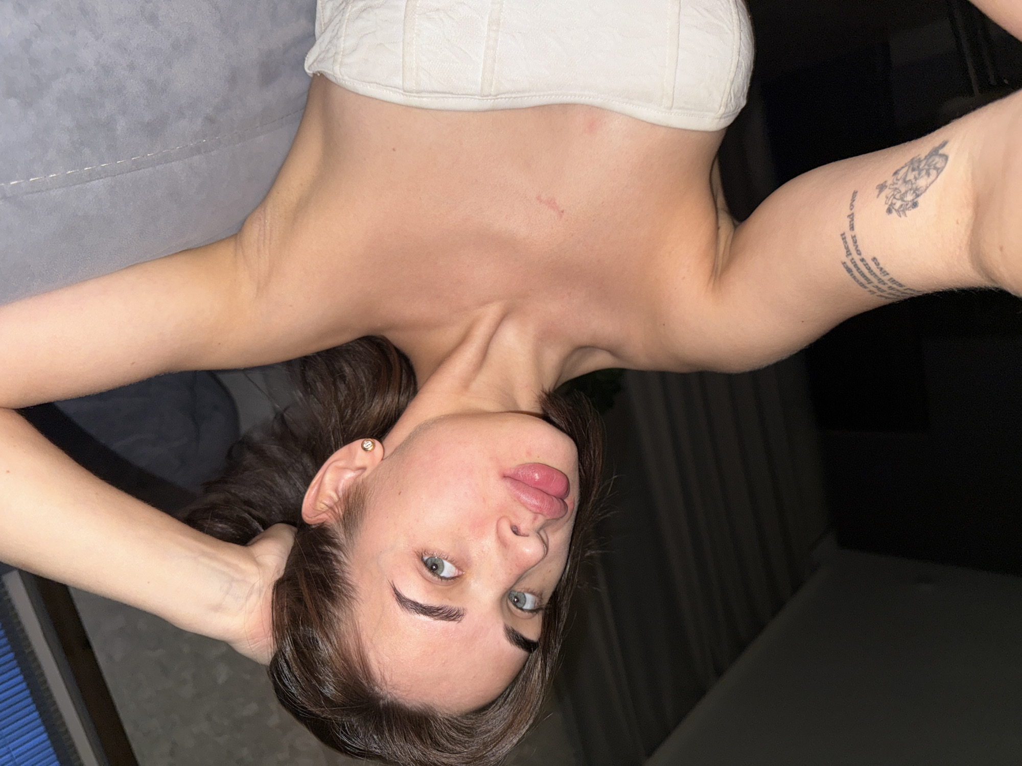 A woman with brown hair and light eyes is lying on a light-colored surface with her head angled towards the bottom of the frame. She is wearing a white strapless top. Her arms are extended, with one hand near her head and the other with a tattoo visible. A portion of a grey sofa is visible in the upper left corner, and a dark area is in the background on the right. It is not possible to determine if this image was taken in Лобаново, Russia.