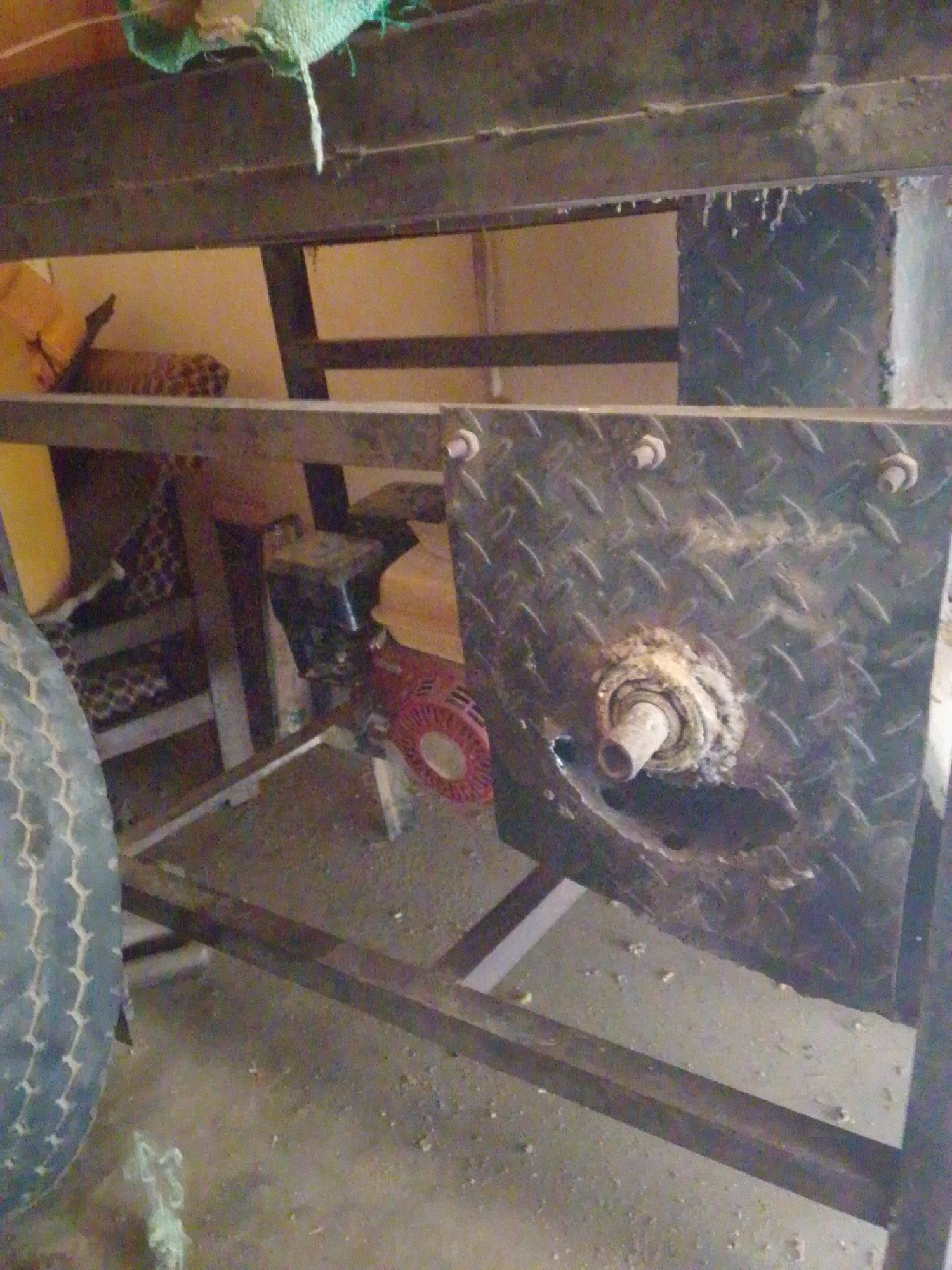 The image displays a close-up of a mechanical apparatus, possibly part of a larger machine, situated indoors. The primary focus is a square, textured metal plate with a prominent central bearing assembly, which appears worn and possibly rusted. This plate is attached to a metal frame. To the left of the plate, a portion of a tire is visible, suggesting the apparatus might be mobile or part of a vehicle-like structure.
Behind the metal plate and frame, a red component with a circular grate is visible, likely a fan or filter, attached to a dark, box-like engine or motor. A patterned fabric or object is also visible in the background, partially obscured by the metalwork. The overall setting appears to be a workshop or storage area, with a concrete floor and hints of a wall in the background. The lighting is dim and appears to be artificial, suggesting an indoor environment, possibly a garage or shed. No people are visible in the image, and there is no discernible text. The scene conveys a sense of utilitarian machinery, possibly in a state of disrepair or under construction, with no specific indication of the time of day, weather, or emotional tone beyond the functional nature of the equipment.