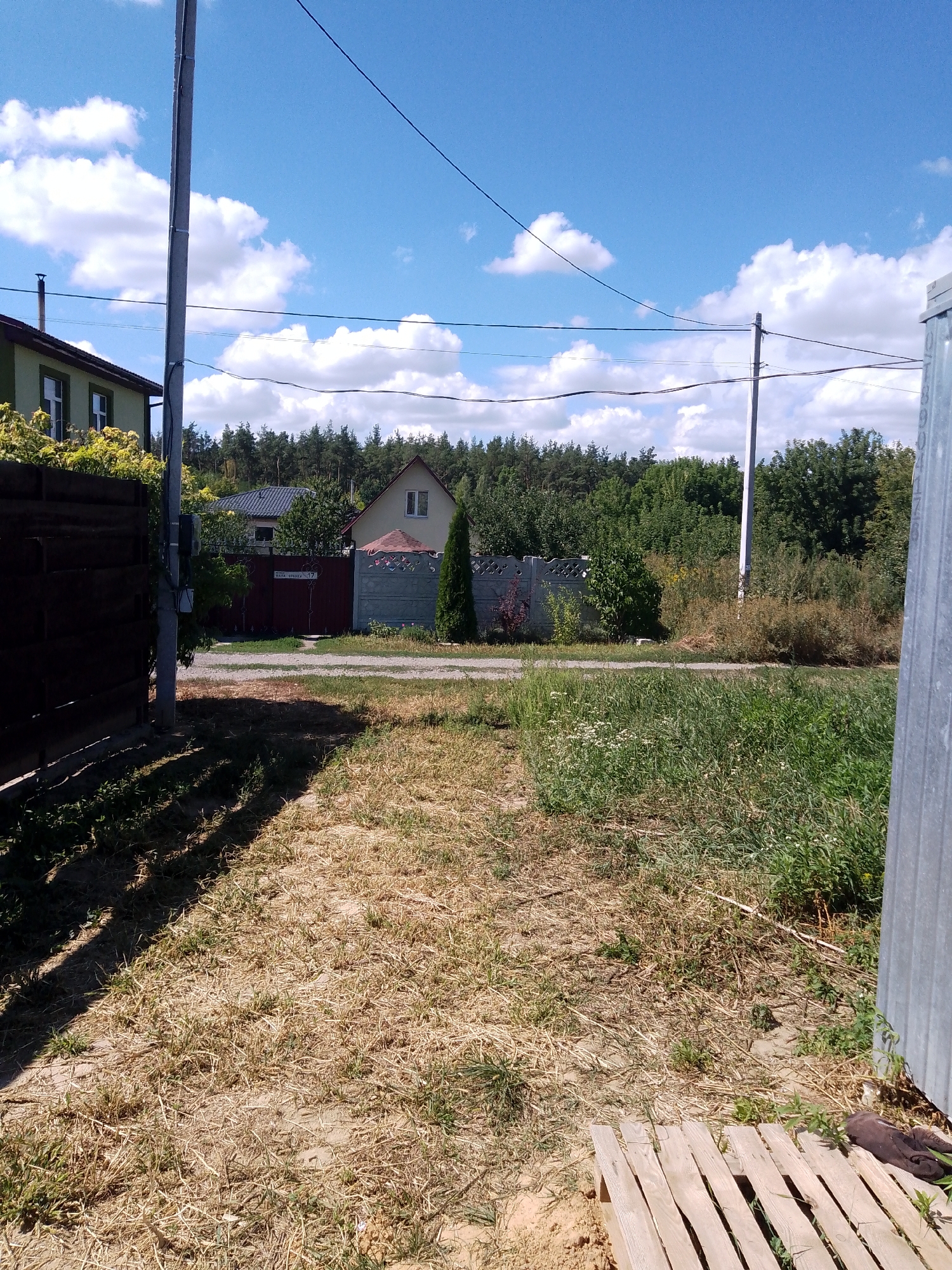 The image shows a rural scene on a sunny day in Zalizne, Ukraine. In the foreground, a dirt path is partially covered with dry grass and scattered green weeds. To the left, a dark wooden fence casts a shadow across the path. In the midground, there is a residential area with houses and fences. A house with a green facade and white trim is visible on the left, behind a dark wooden fence. A lighter-colored house with a beige roof and a small gable window is situated further back. Between the houses are various fences, including a dark metal gate with a sign and a decorative concrete fence. A tall, thin evergreen tree stands next to the concrete fence. The background features a dense forest of coniferous trees. Several utility poles with wires are present, with one prominent pole in the mid-right of the image. The sky is bright blue with scattered white cumulus clouds. To the far right, a portion of a corrugated metal structure is visible.