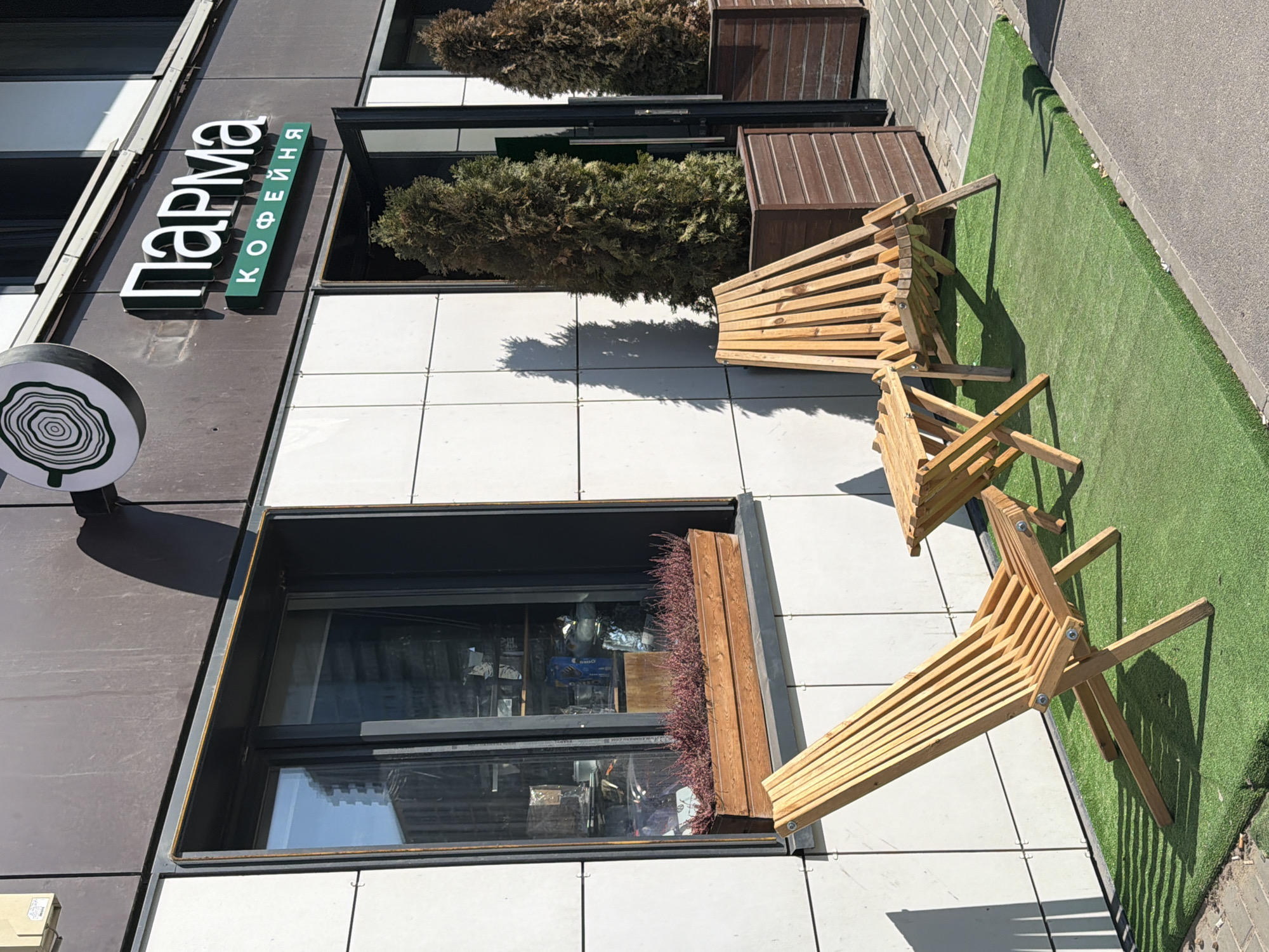 The image captures an outdoor seating area outside a cafe in Saint Petersburg, Russia. The cafe's facade features a prominent sign with "Парма КОФЕЙНЯ" in white and green lettering. The building's exterior is clad in a combination of dark grey and white tiled panels.
In the foreground, two modern wooden chairs are set up on a patch of artificial green turf, which is bordered by a paved area and an asphalt road. The chairs, made of light-colored wood, have a slatted design and are angled towards each other, suggesting they are meant for patrons to relax. The sun casts distinct shadows from the chairs and nearby plants onto the white tiled surface, indicating it is daytime and likely sunny.
Behind the chairs, a planter box with a cluster of small, reddish-brown plants sits in a window recess of the building. Further back, two large evergreen trees in wooden planters add to the greenery of the outdoor space. The overall scene suggests a pleasant and inviting atmosphere for a cafe.
There are no people visible in the image. The setting appears to be a commercial establishment, possibly a cafe or restaurant, designed to attract customers with its modern aesthetic and comfortable outdoor seating. The architectural style and the presence of the Russian text on the sign confirm the location context.