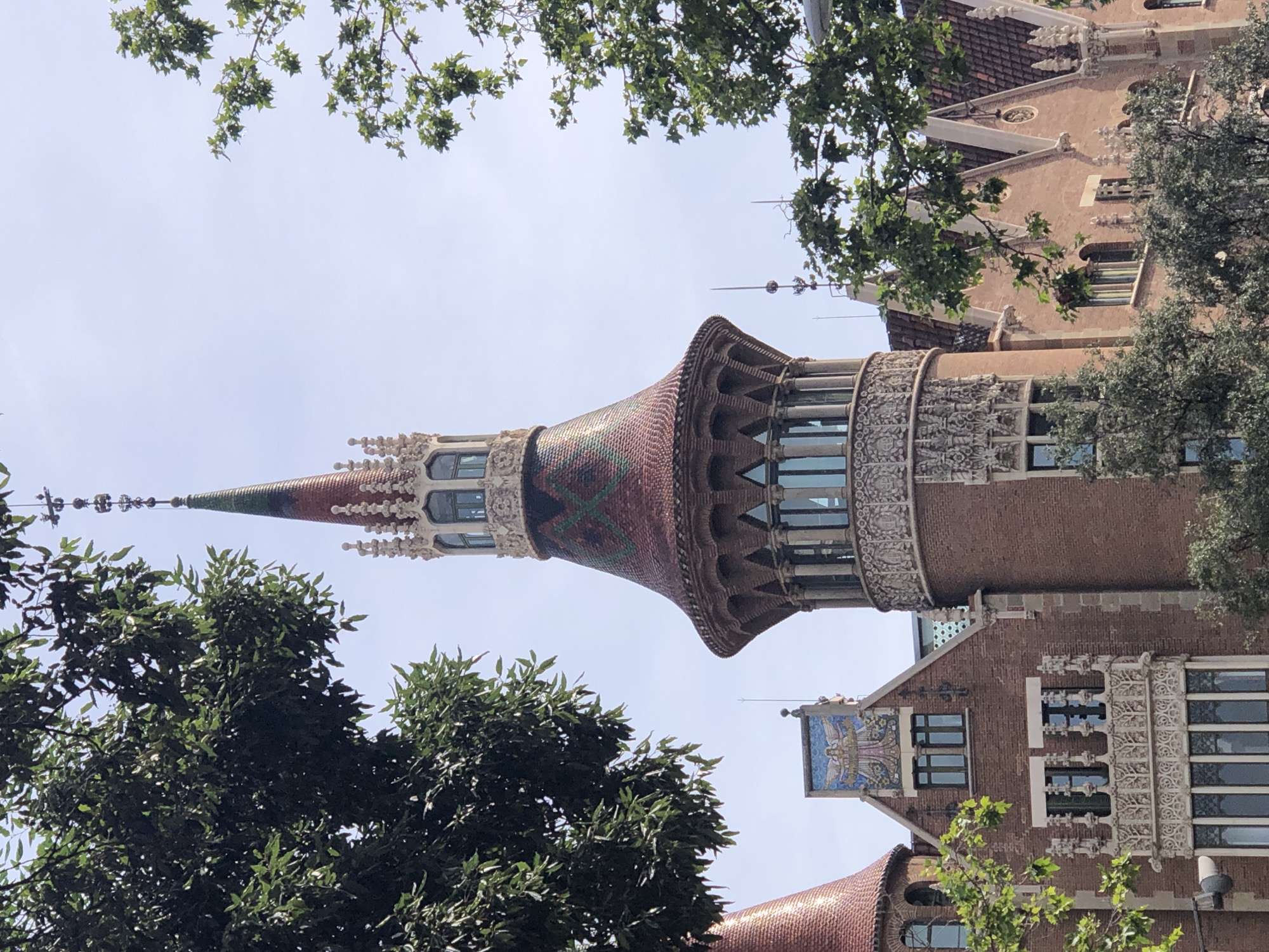 This is a medium shot, eye-level, outdoor, daytime photograph of a historic building with ornate architecture, framed by trees. The building is constructed primarily of red brick and features a prominent tower with a conical, tiled roof. The roof tiles are a mix of red and green, arranged in a diamond pattern. The tower has decorative stonework and arched windows. Adjacent to the tower are other sections of the building with tiled roofs, gabled sections, and elaborate window detailing, including a mosaic of a winged figure above a prominent window. The sky is a clear, pale blue. Lush green foliage from trees overhangs the scene from the left and right sides, partially obscuring the building. The overall impression is of a well-preserved, architecturally significant structure in a verdant setting.